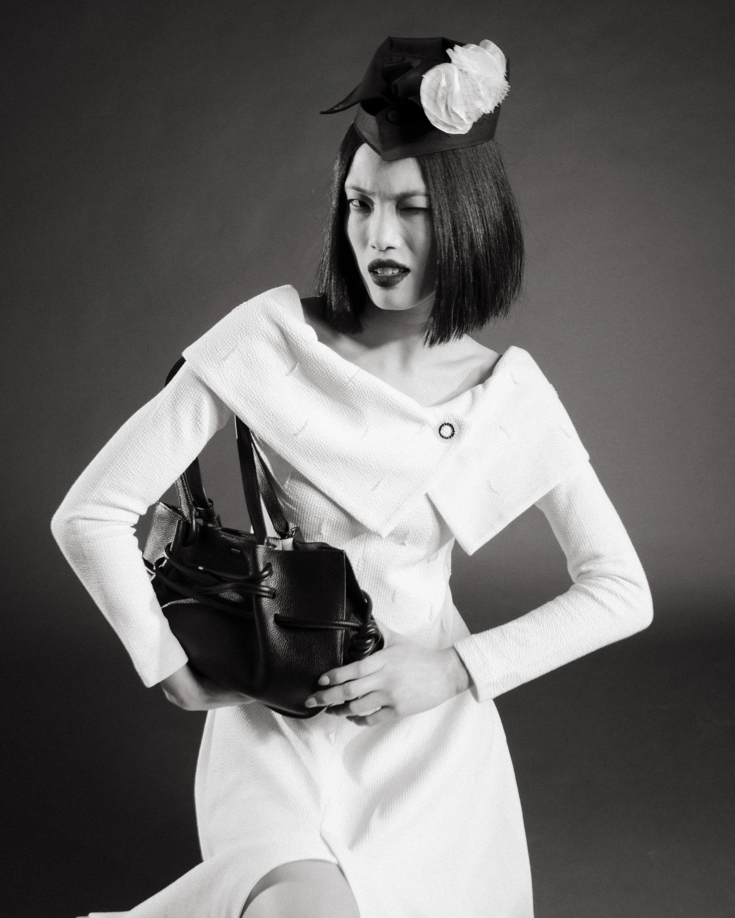 A black-and-white photo of a woman with a short bob haircut, wearing a white dress or coat with a textured fabric. She has dark lipstick and is wearing a small hat adorned with a large flower on top. She appears to be winking or squinting, with a playful expression. She is holding a large, glossy black handbag on her left shoulder, with her left hand resting on her hip and her right arm crossing in front of her. The background is plain and dark.