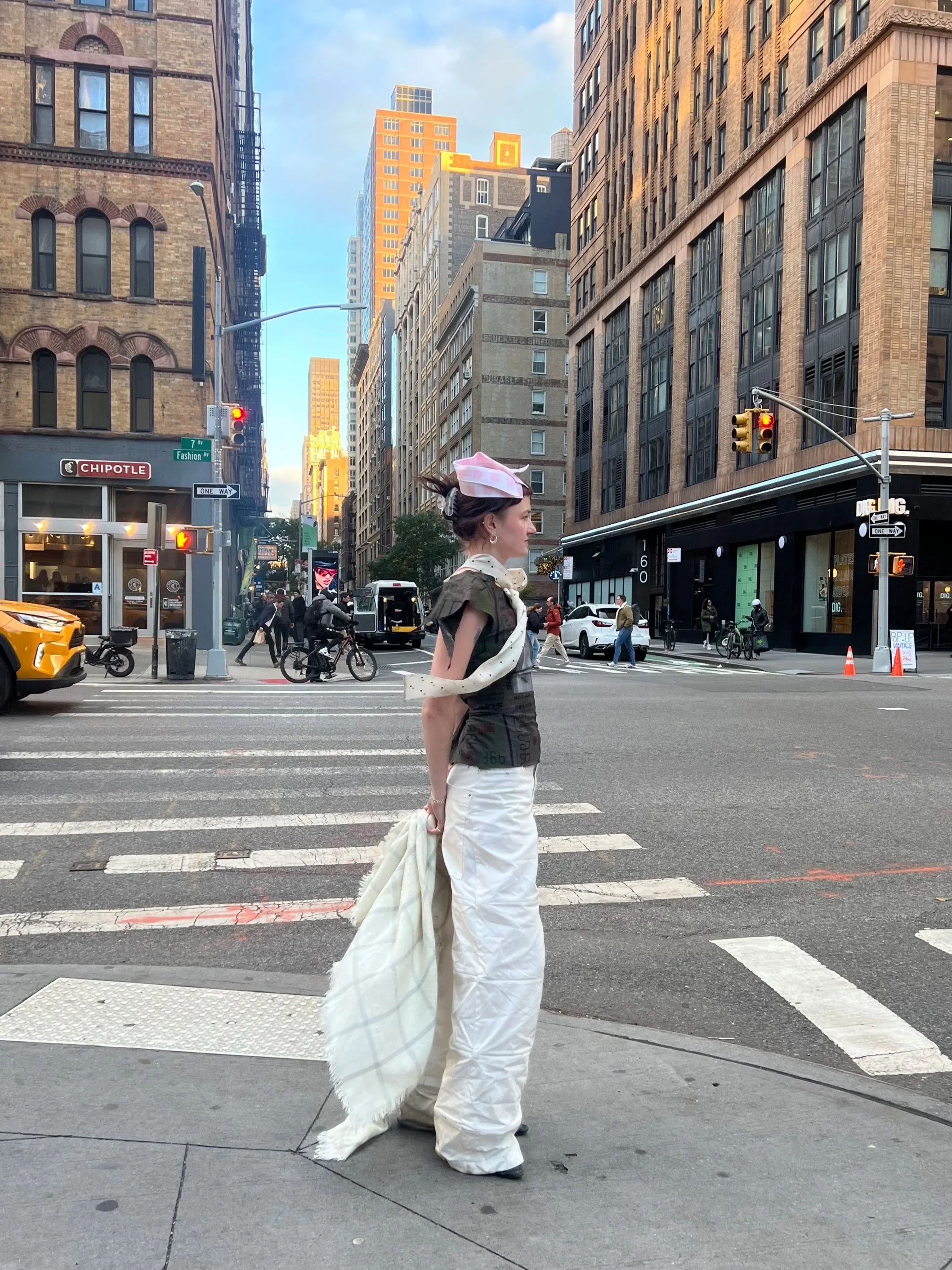 Fashionable woman crossing the street in a city with high-rise buildings, traffic lights, and pedestrians, wearing a headscarf, a patterned jacket, and white wide-leg pants.