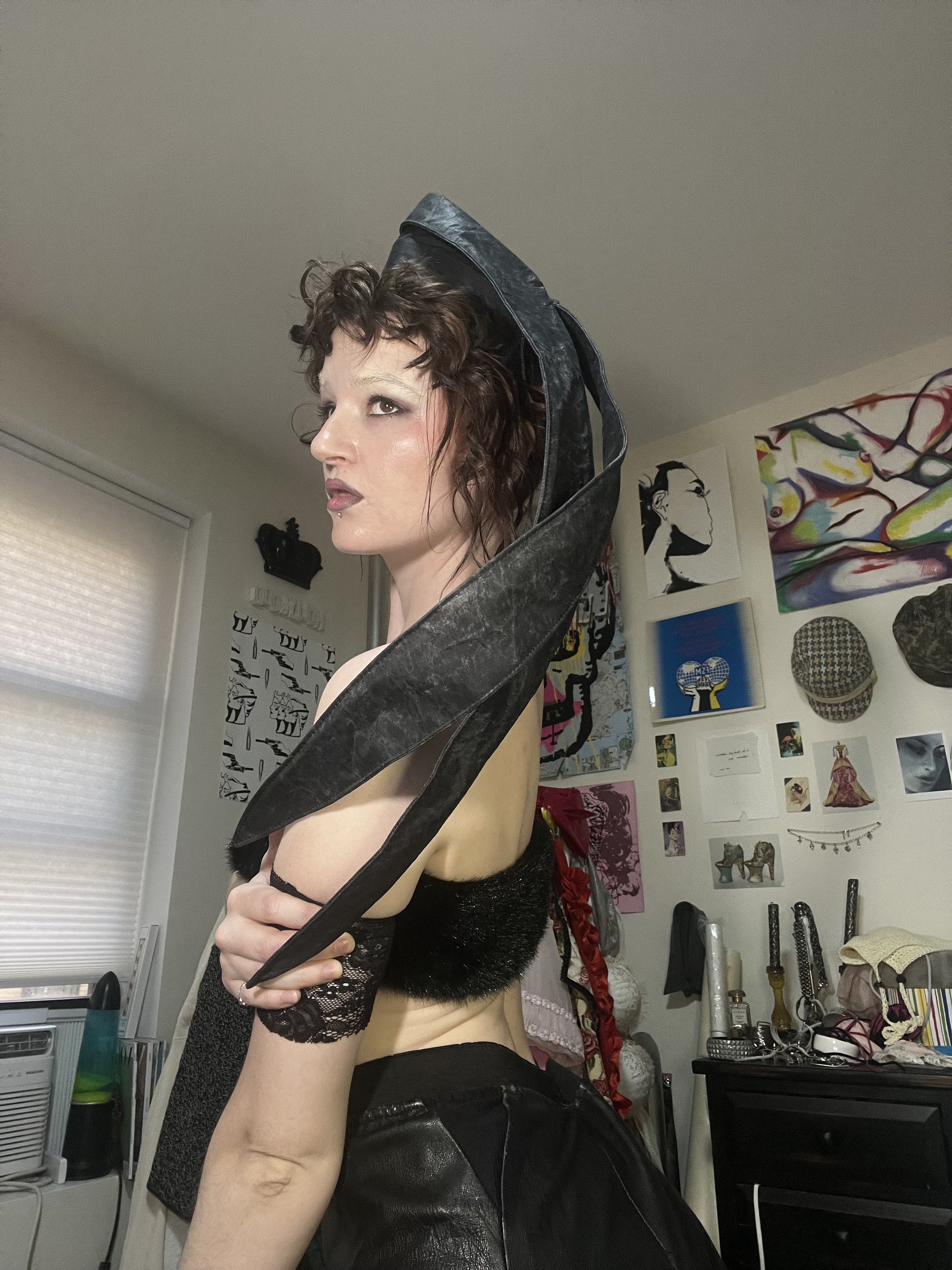 A woman with short curly hair and makeup, wearing a large black leather collar; she is looking sideways and is partially dressed in black lace clothing, standing in a room with colorful artwork and various items in the background.