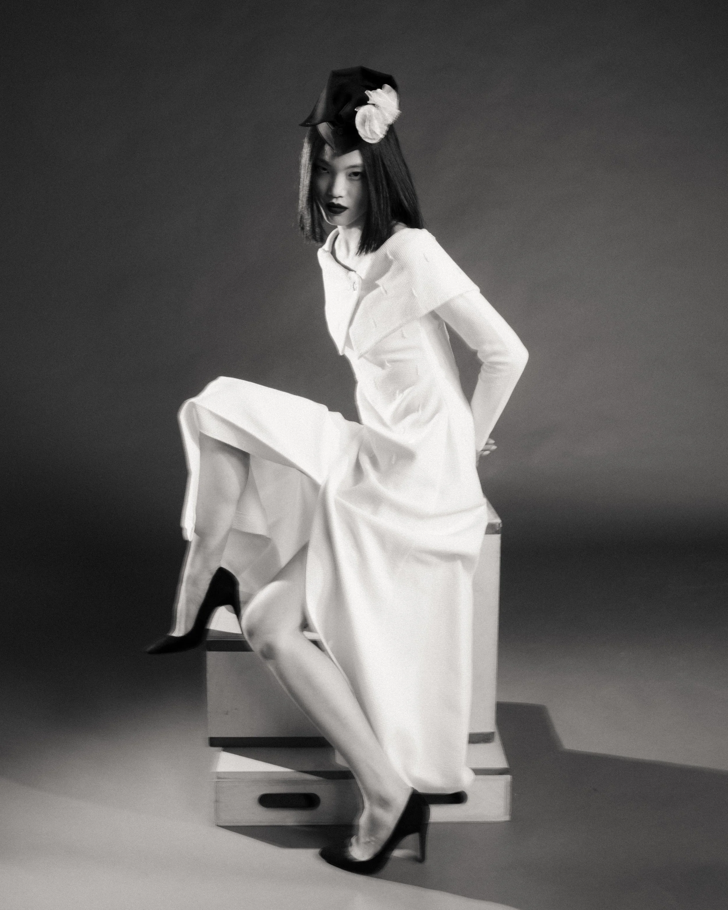 A black and white photograph of a woman sitting on a chair with a plain gradient background. She has dark, straight hair and is wearing dark lipstick. She is dressed in a white outfit with a large collar, and is wearing black high heels. She is looking at the camera with a confident expression and has one arm resting behind her.