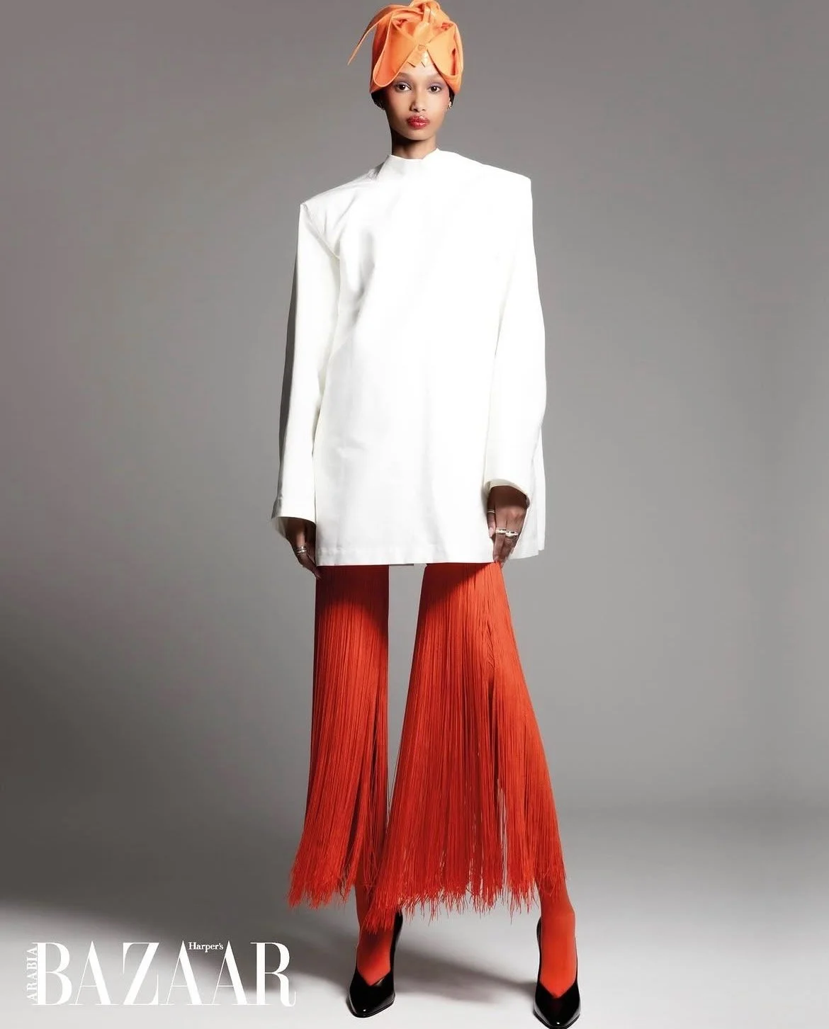 Model wearing a white high-neck coat, orange pleated wide-leg pants, orange head wrap, and black and orange heels, standing against a gray background, as part of a fashion photoshoot for Harper's Bazaar.