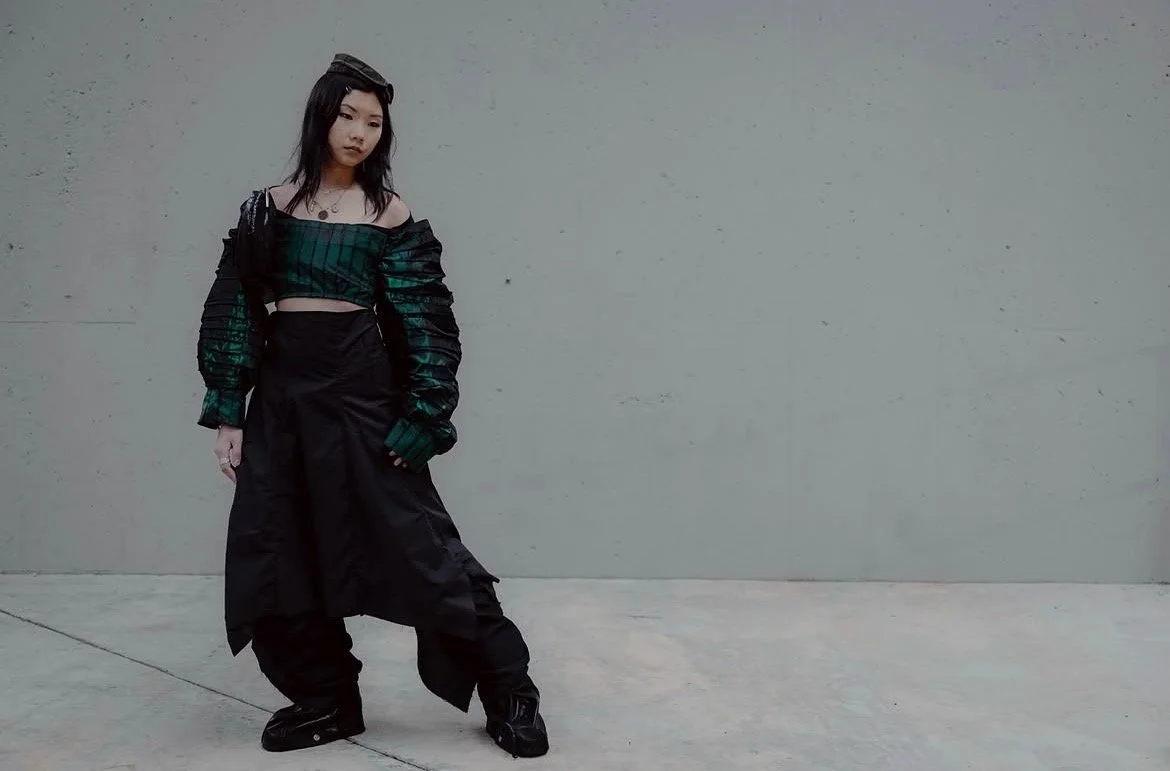 A young woman with black hair and a headband, dressed in a black and green off-the-shoulder crop top, black baggy pants, and black shoes, standing against a plain gray wall.