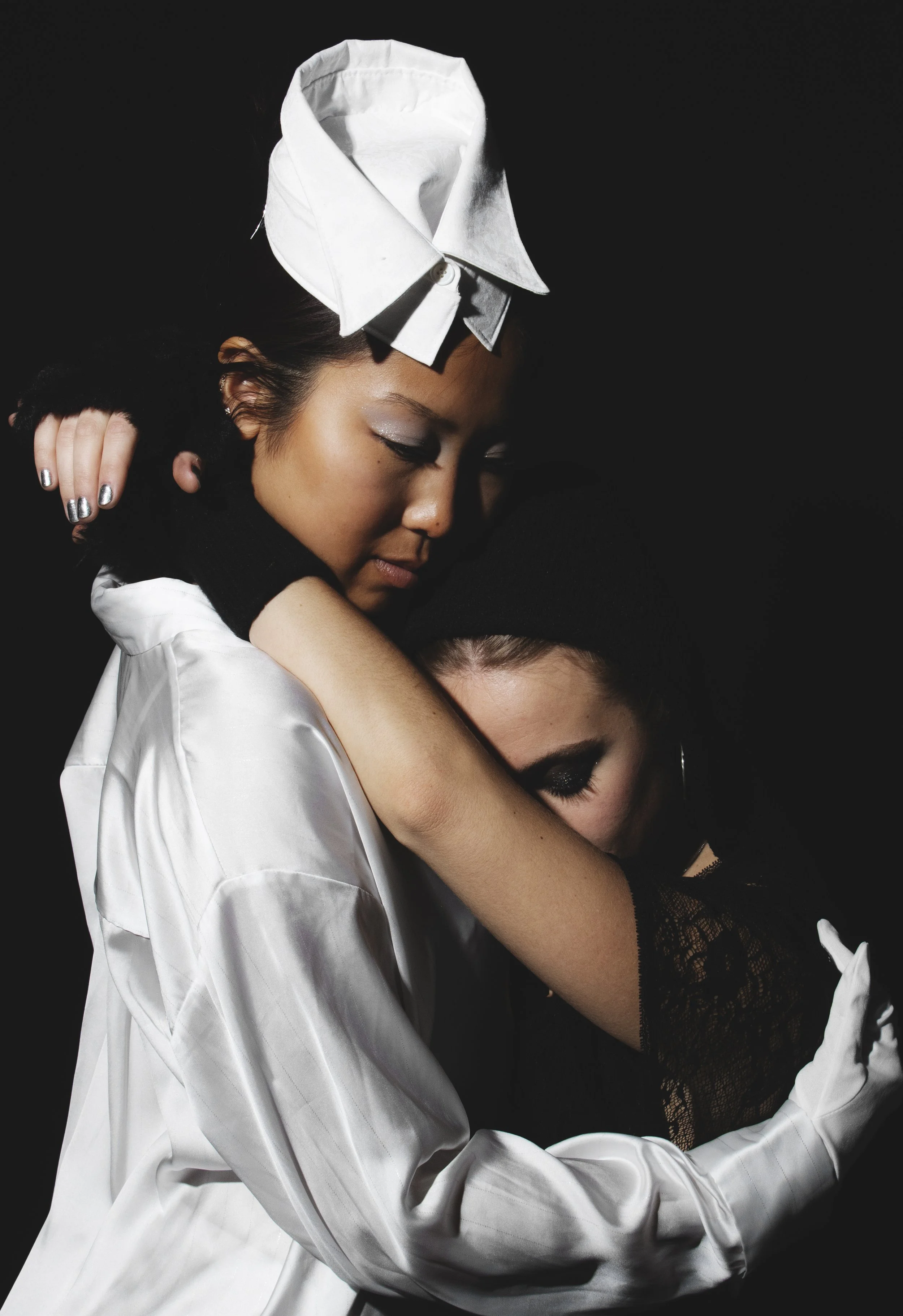 Two women hugging, one wearing a white satin shirt with a white headpiece, the other wearing black lace gloves, makeup, and a black top, against a black background.