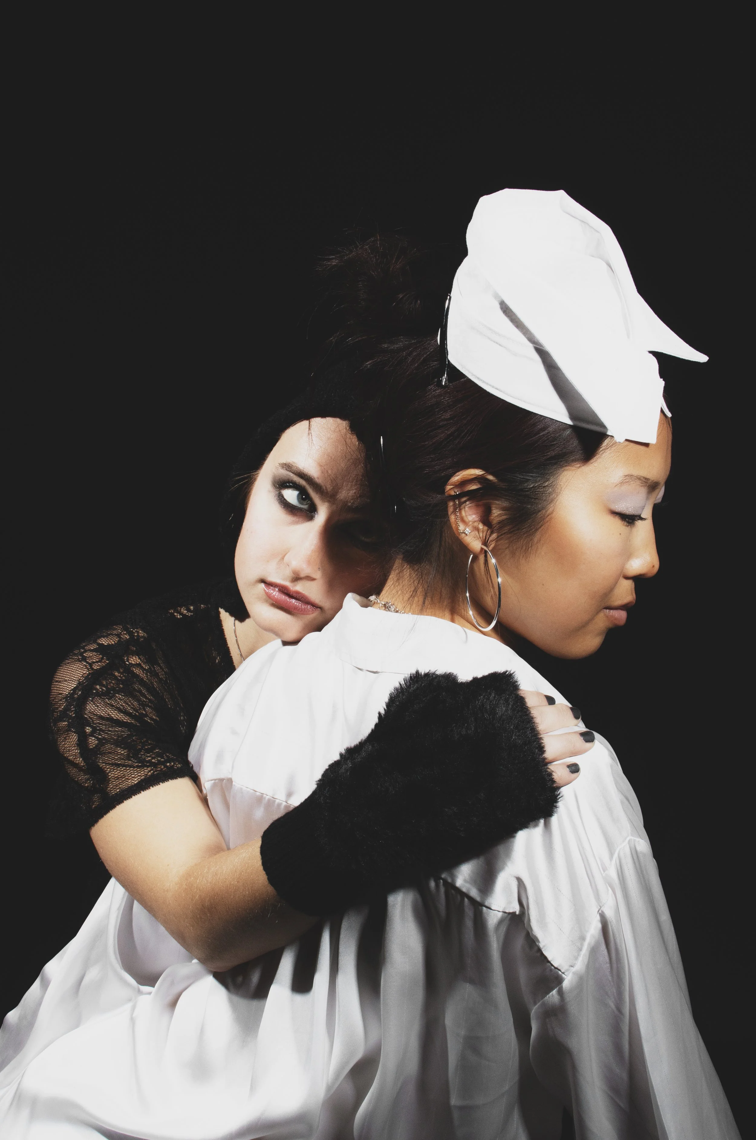 Two women embracing against a dark background. One woman has dark hair and tattoos, wearing a black lace top and black gloves. The other woman has brown hair, closed eyes, and earrings, wearing a white headscarf and satin outfit.