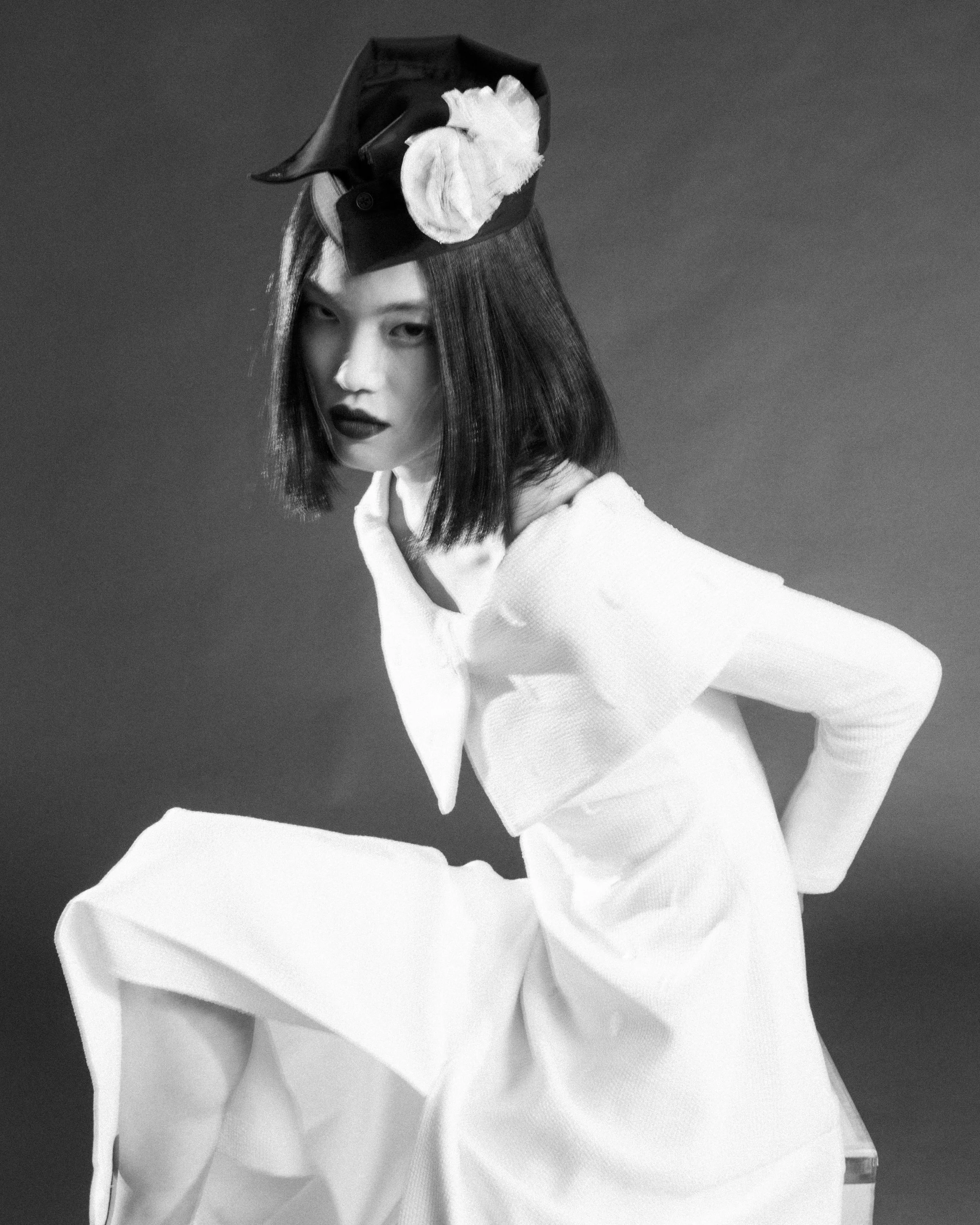 Black and white photo of a woman with chin-length straight hair, wearing a white textured garment, and a headpiece with large fabric flowers.