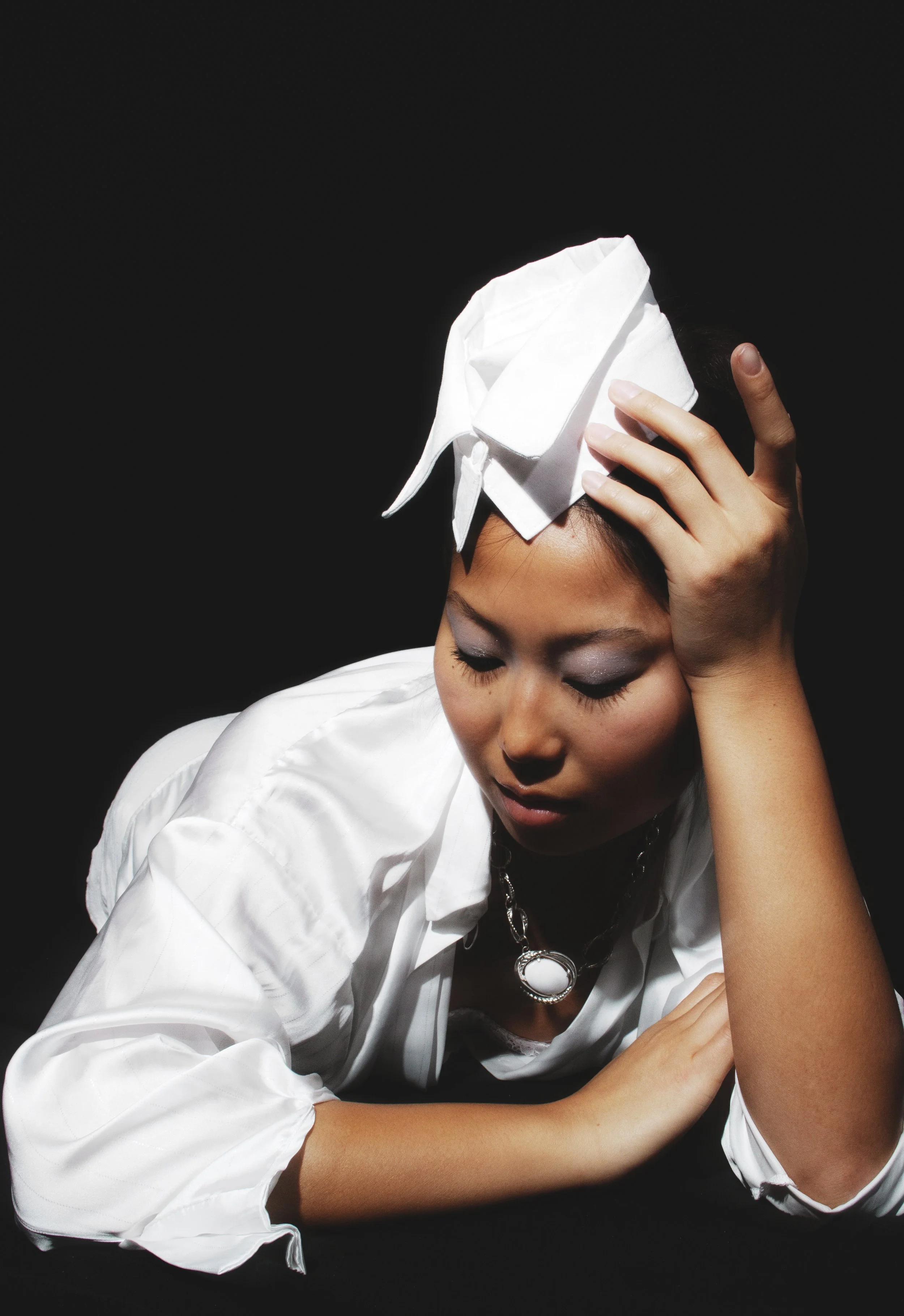 A woman wearing a white satin blouse, a large white bow on her head, silver necklaces, and makeup, resting her head on her hand against a black background.