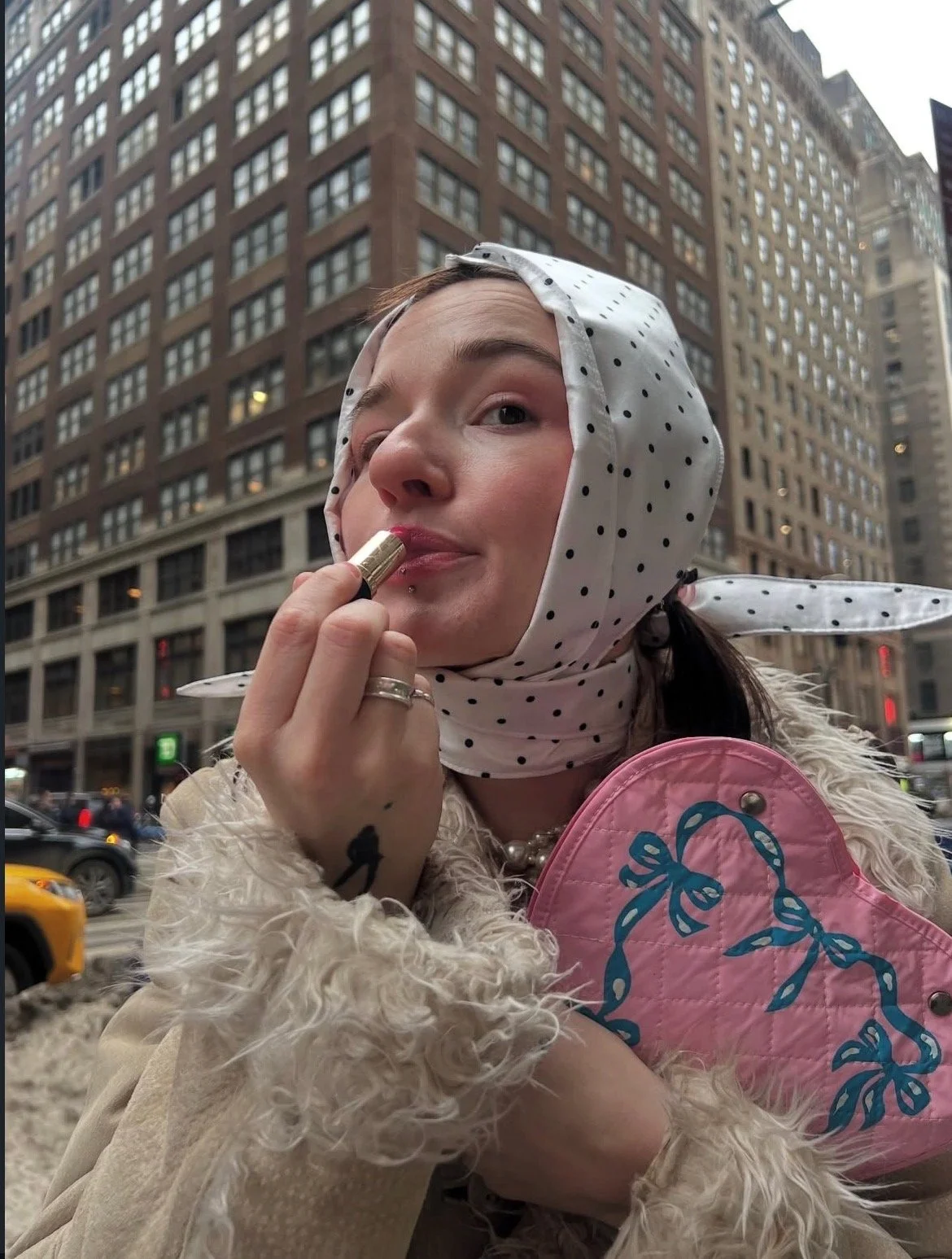 A woman in a polka dot headscarf and fluffy beige coat is looking slightly upward while applying lipstick, standing on a city street with tall buildings and traffic in the background.