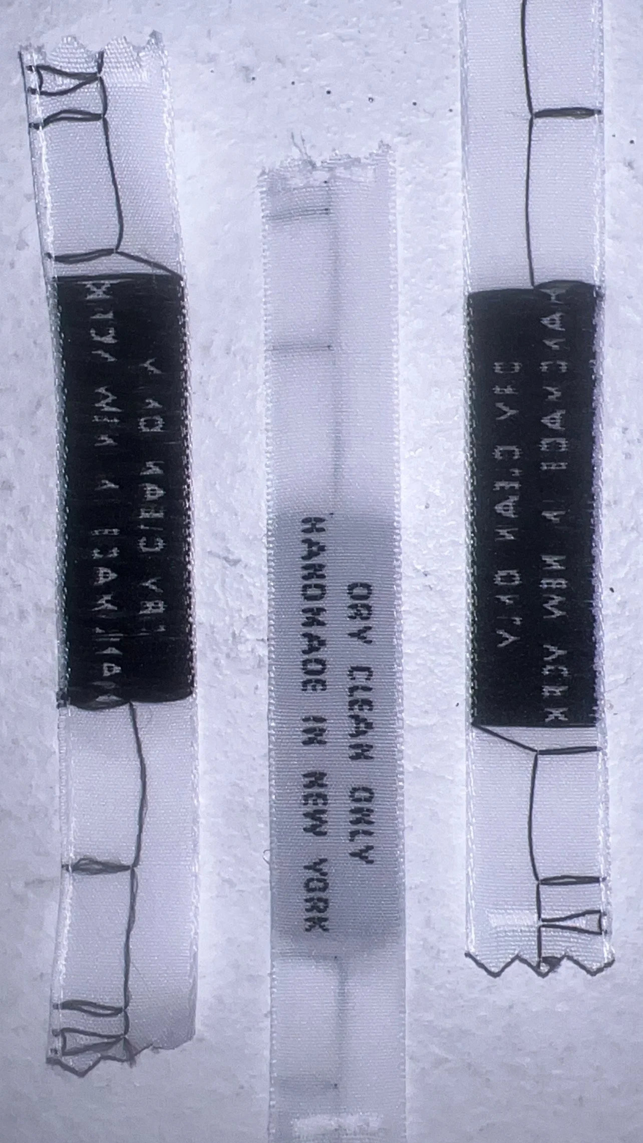 Three stitched fabric labels with black and white text, two on the left and right, and one in the center. The labels are attached to a white textured background with visible stitching.