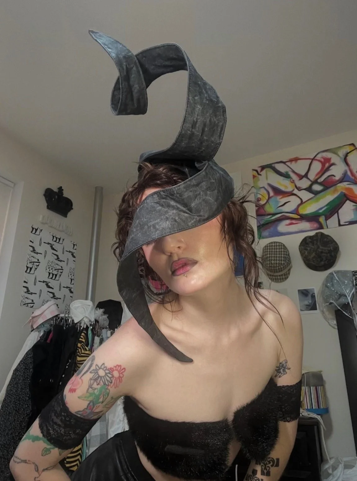 A woman with tattoos and wavy hair wearing a black lace top, with a large black and grey spiral sculpture wrapped around her head in a room with abstract art on the walls and hats on display.