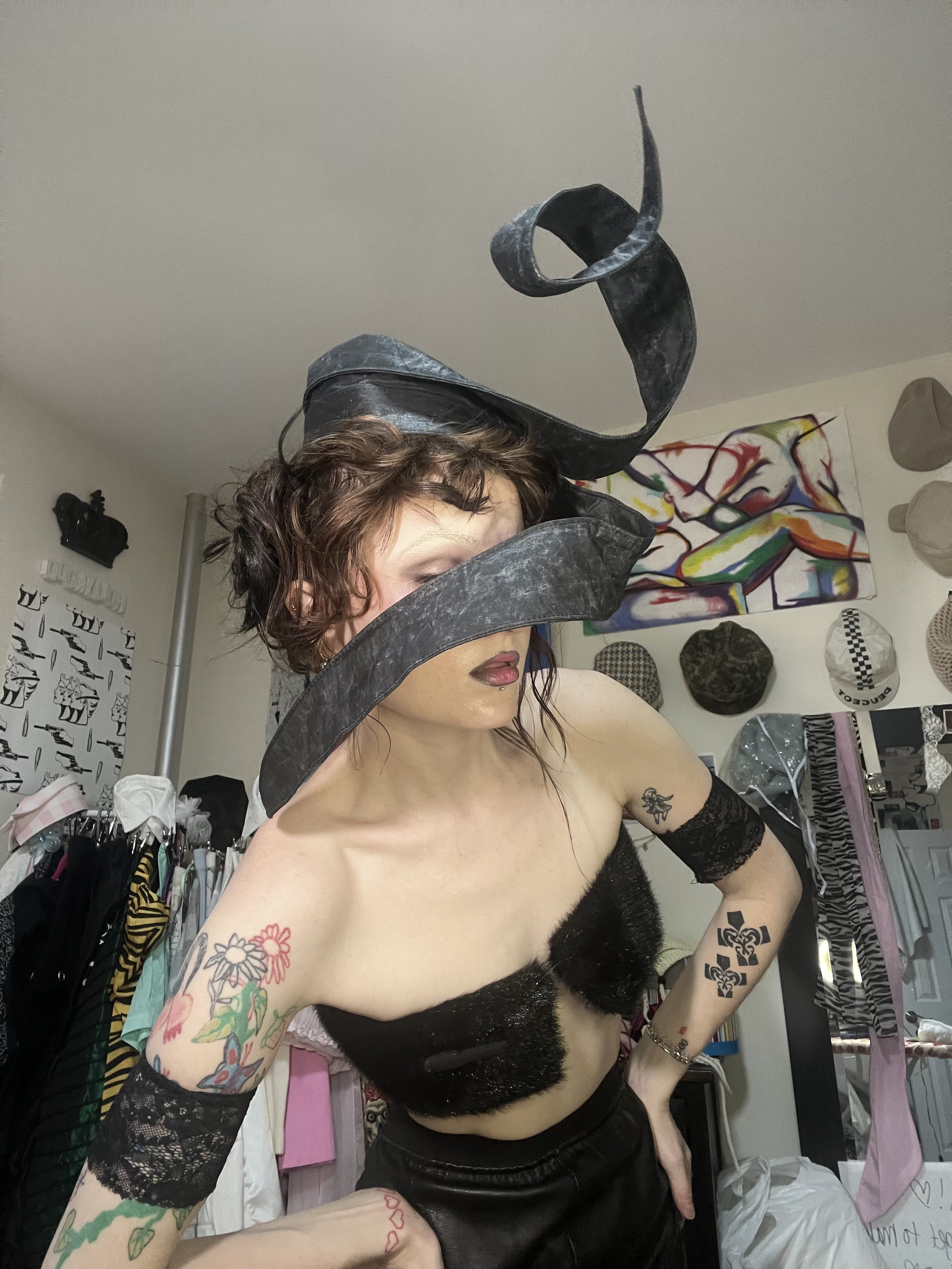 A woman with short curly hair and multiple tattoos, wearing a black lace arm and a black fuzzy top, poses in a room with colorful artwork and various items on the walls and shelves.