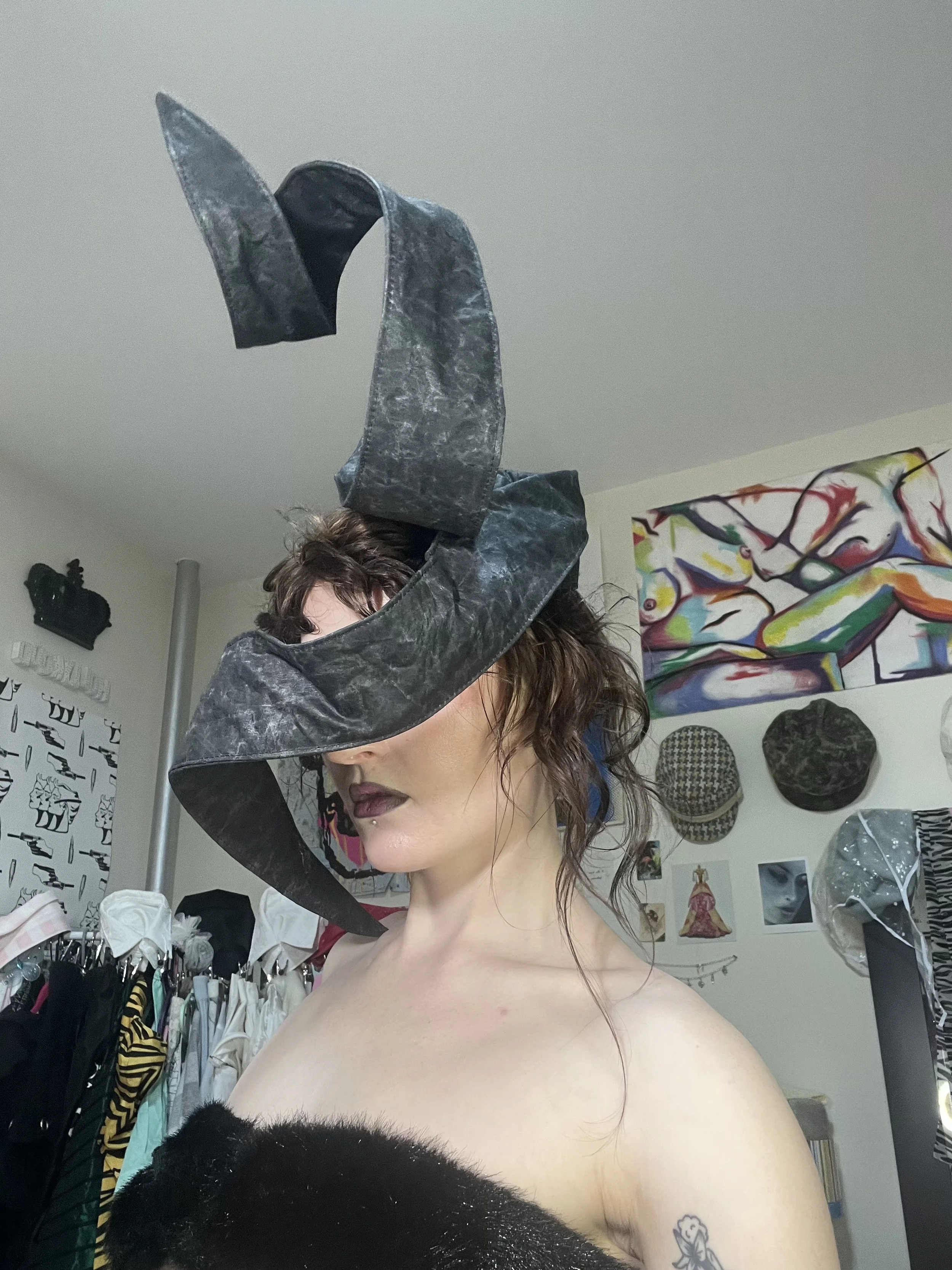A person with on makeup, tattoo, and curly hair, wearing a black headpiece with long, curved, draped fabric. The background features artwork, hats, and clothing in a room resembling an art studio or boutique.