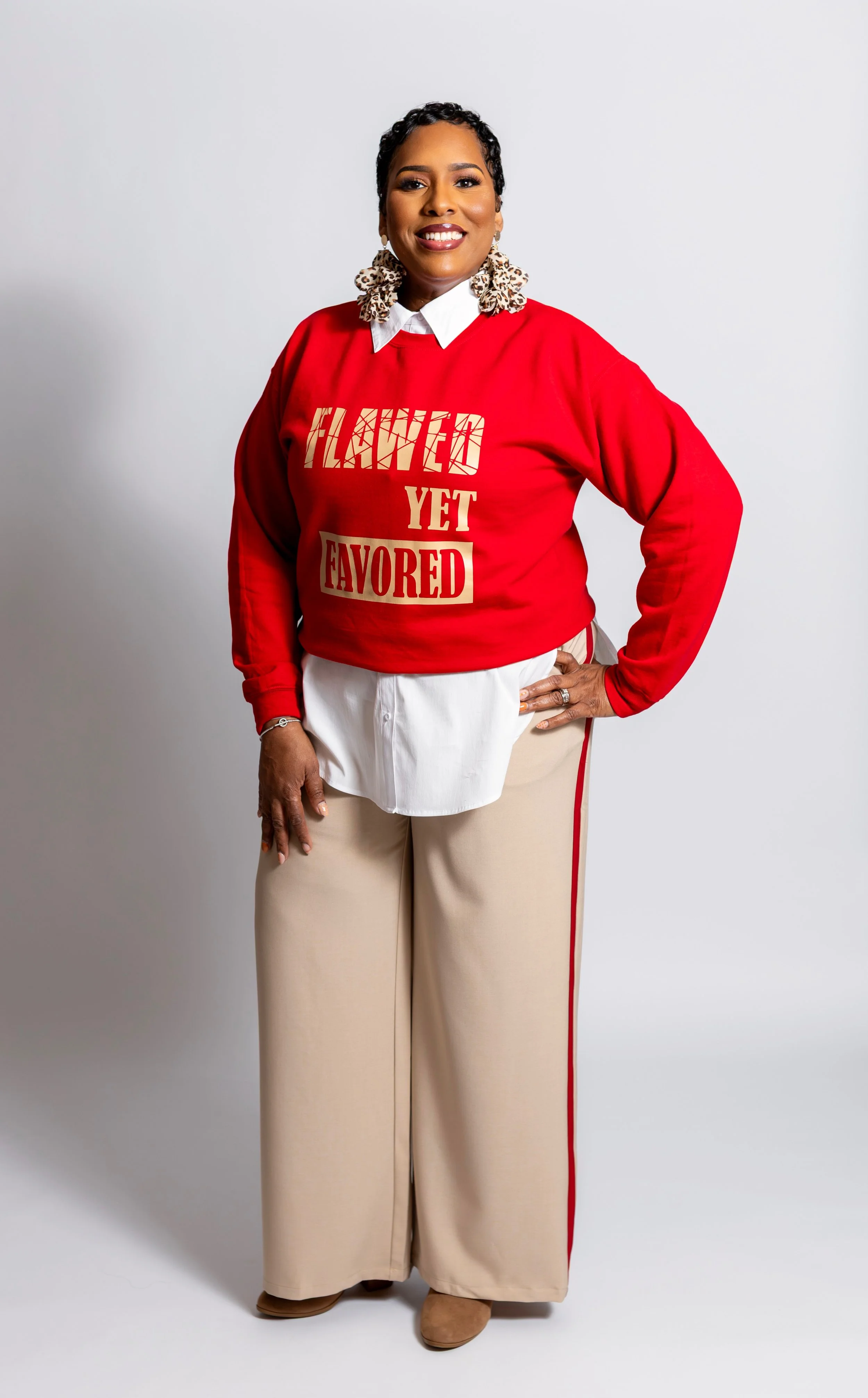 Woman with short curly hair smiling, wearing a red sweatshirt with the text 'HAVED YET FAVED' in gold and beige, white shirt underneath, beige wide-leg pants with a red stripe, and leopard print earrings, standing against a plain white background.