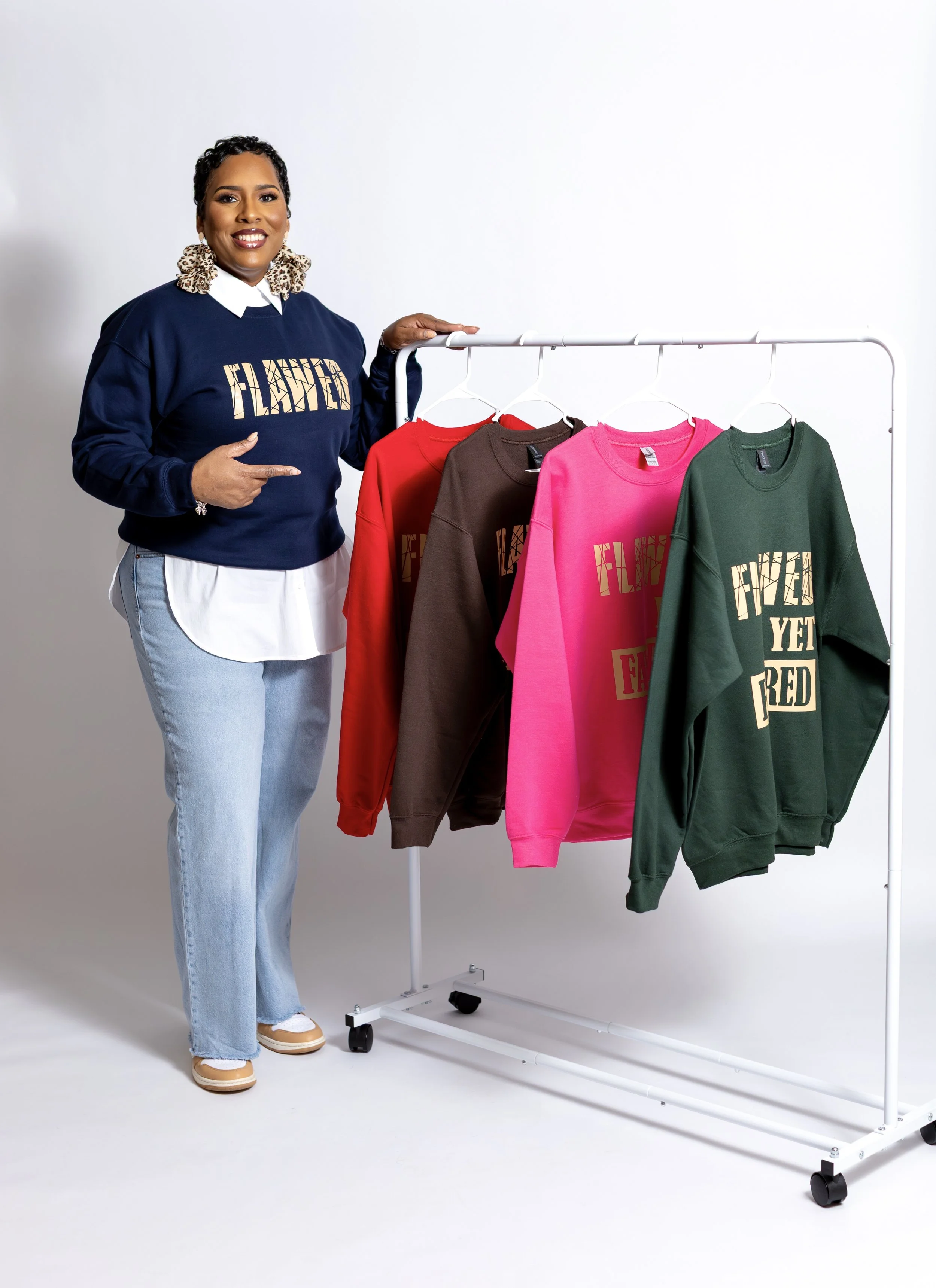 Woman smiling and pointing at a clothing rack with colorful sweatshirts in red, brown, pink, and green.