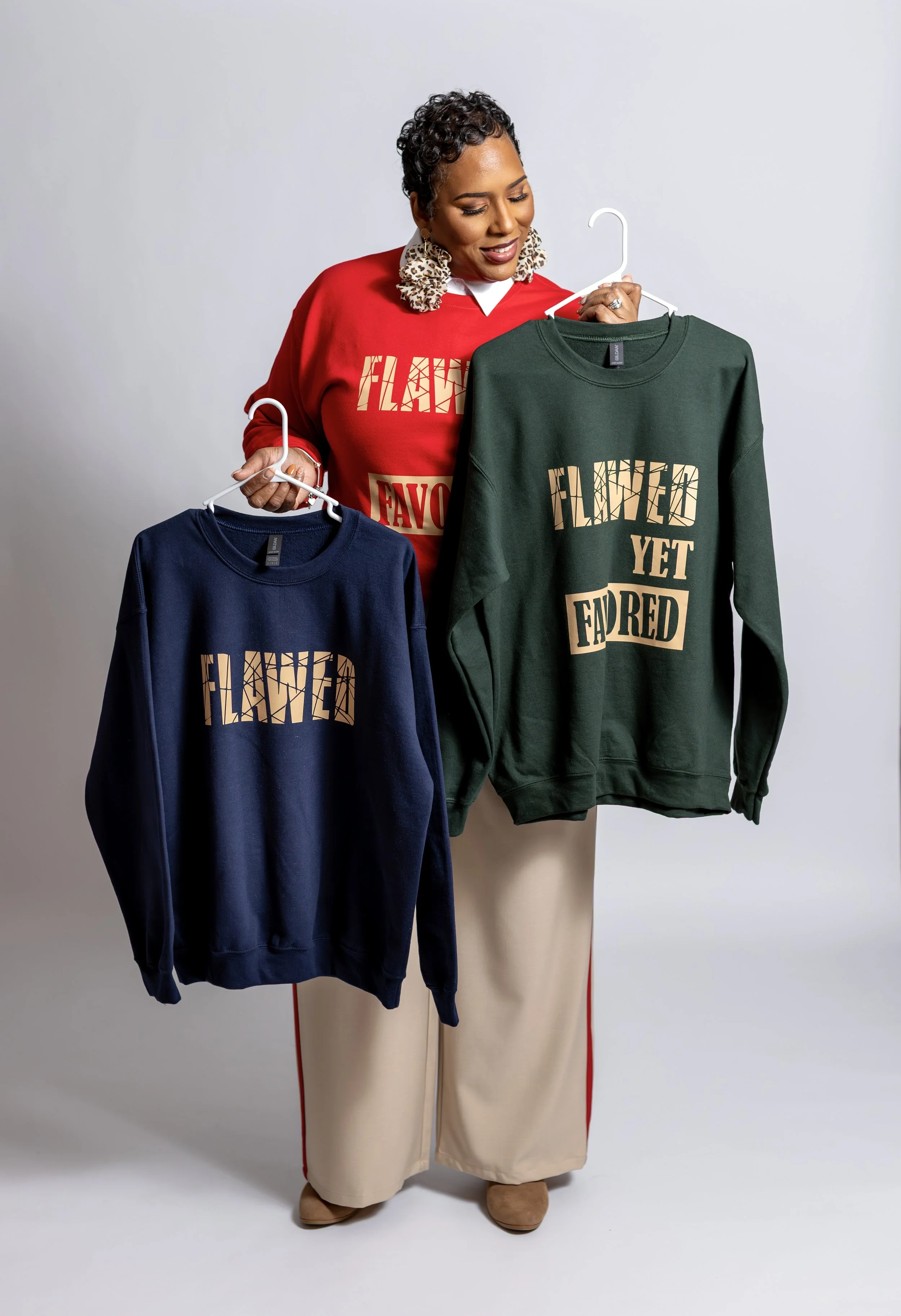 A woman with short, curly hair and leopard print earrings is holding and looking at three sweatshirts on hangers that say 'Flawed Yet Faved' in a photo studio with a plain background. She is wearing a red sweatshirt with the same phrase.