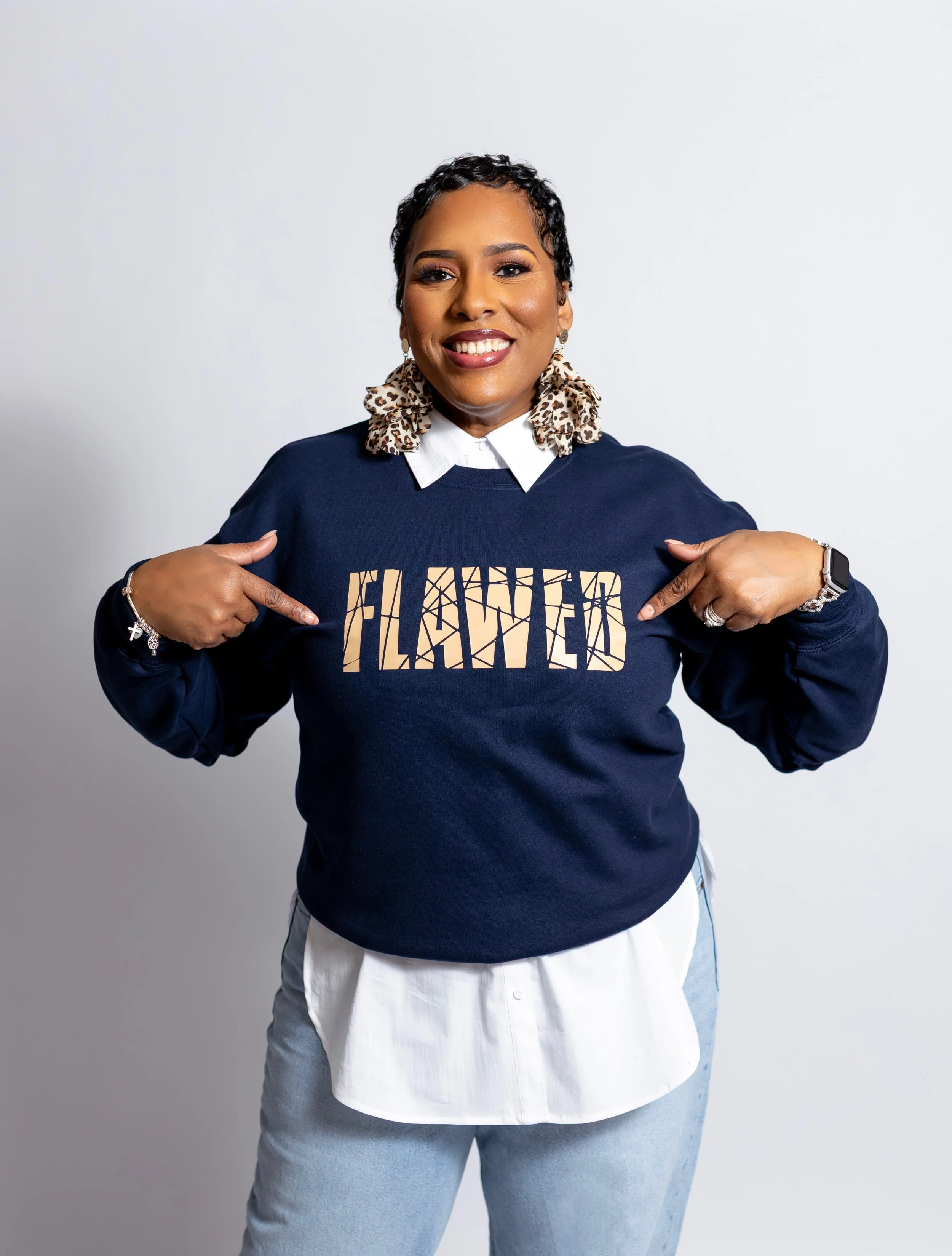 A woman with dark skin and short black hair smiling while pointing to her sweatshirt that reads "I PLANNED." She wears a navy sweatshirt with leopard print scrunchies, earrings, bracelets, and a smartwatch, standing against a plain white background.