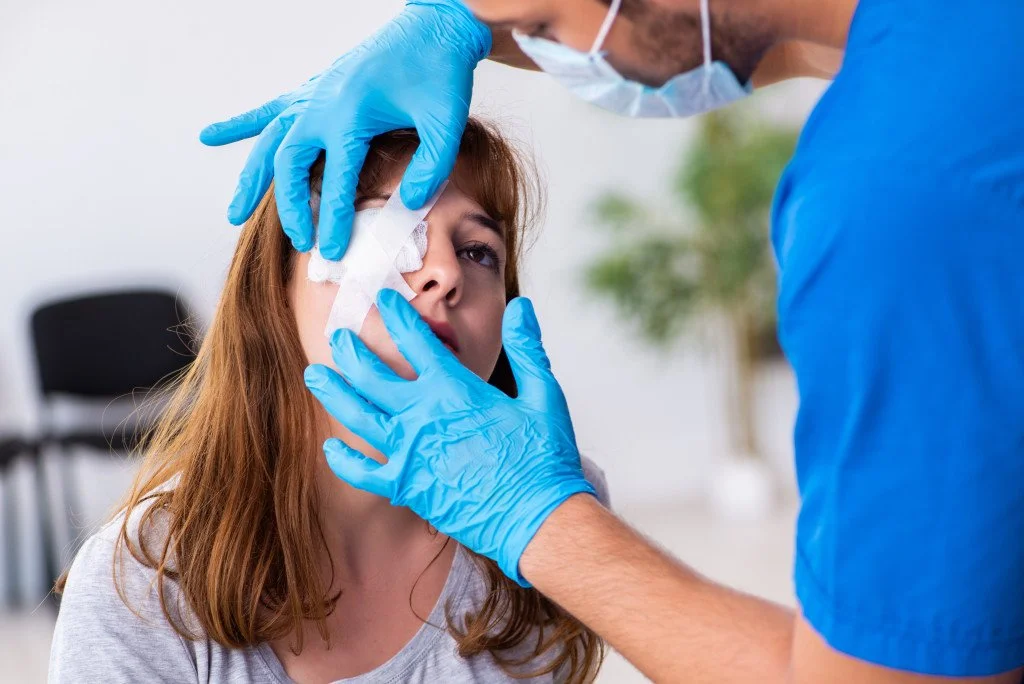 A healthcare professional in blue scrubs and gloves examining a woman's face with a gauze pad on her nose, inside a medical setting.