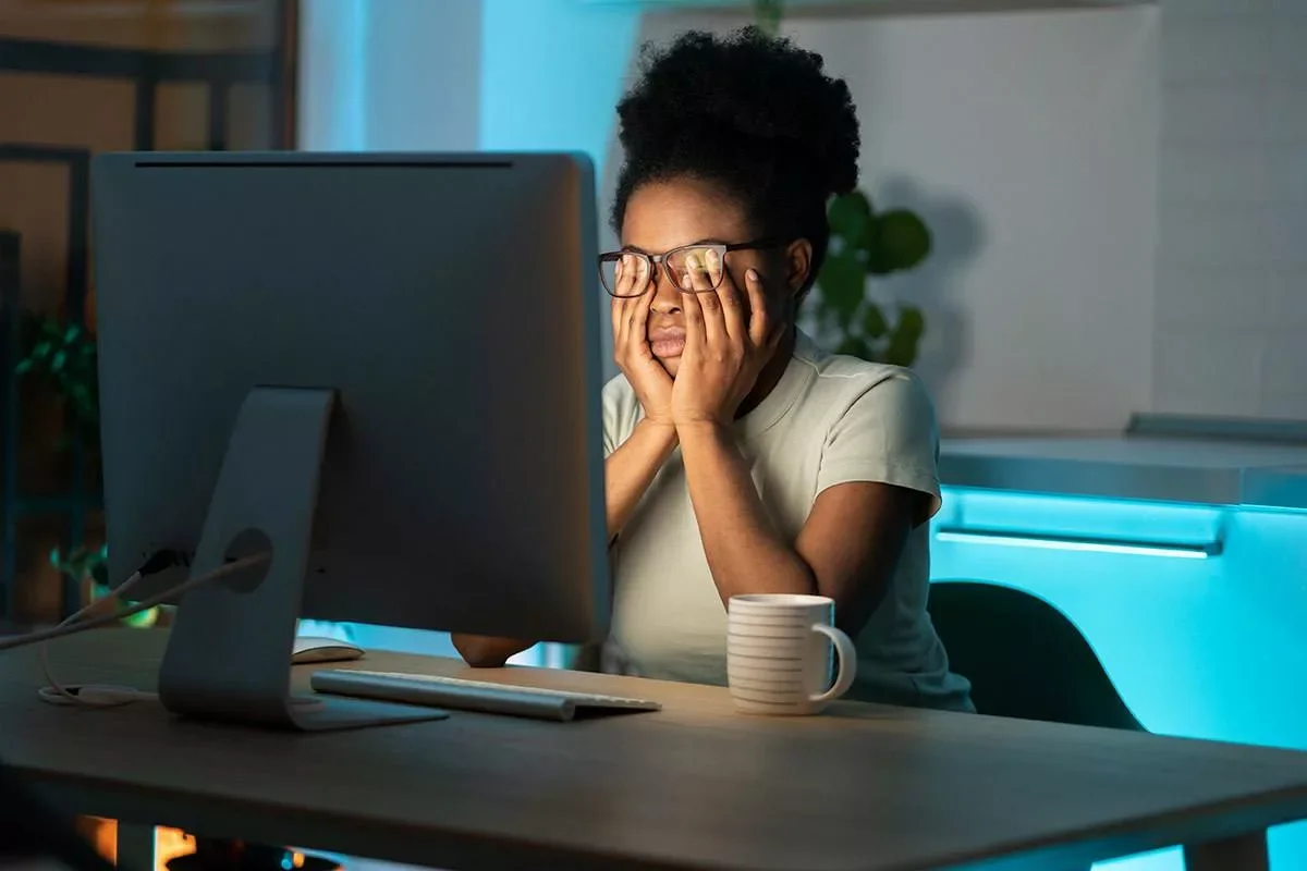 A woman sitting at a desk looking frustrated while working on a computer, with her hands on her face and eyes closed, in a dimly lit room with a mug in front of her.