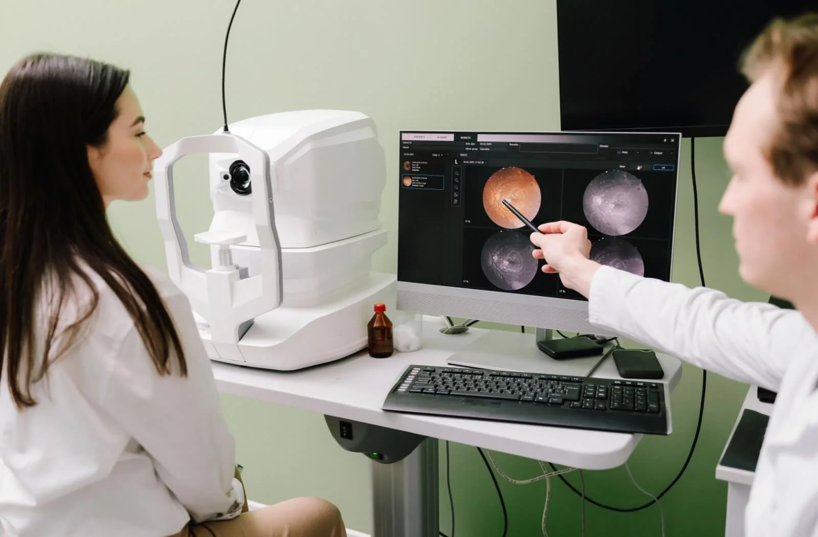 A female patient seated at an eye doctor’s appointment with an eye examination machine on her left and a computer monitor displaying images of her retina on the right, with a doctor pointing at the screen.