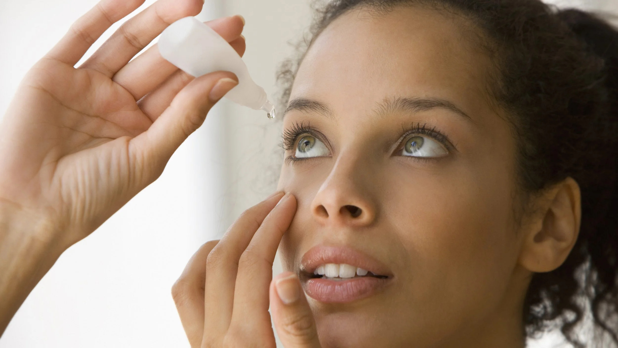 A woman applying a beauty product or skincare serum to her face with a dropper, focusing on her eye and cheek.