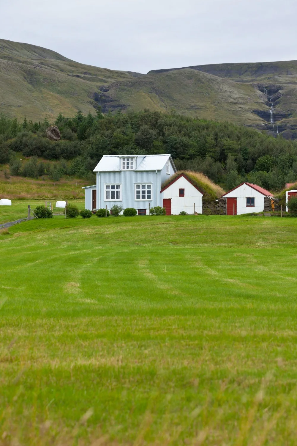 A rural scene featuring a white house with a metal roof, surrounded by smaller white buildings with red doors, set against a lush green landscape with rolling hills and a mountain backdrop.