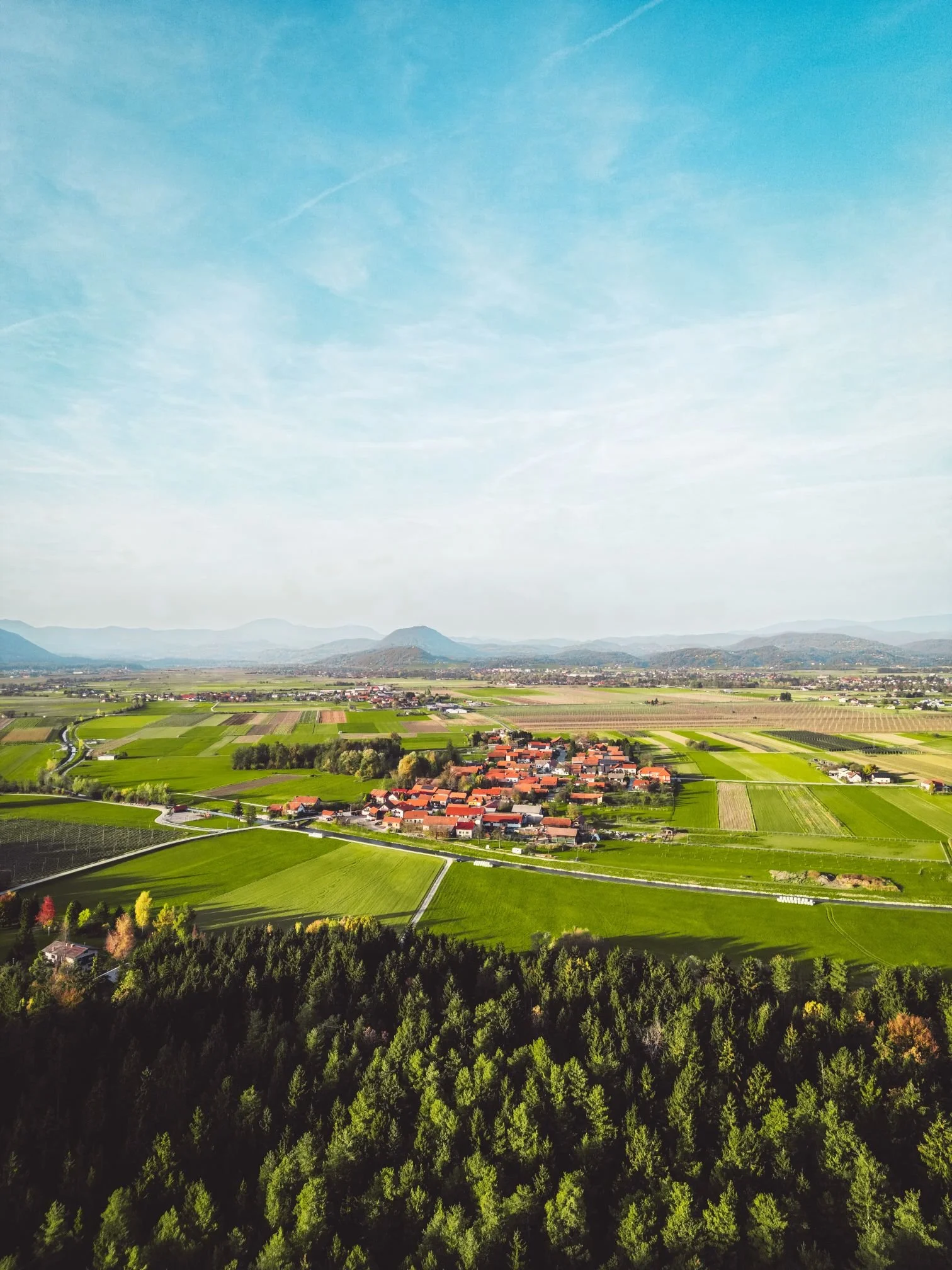 Aerial view of a rural landscape with green fields, a small village with red roofs, and distant mountains under a blue sky.