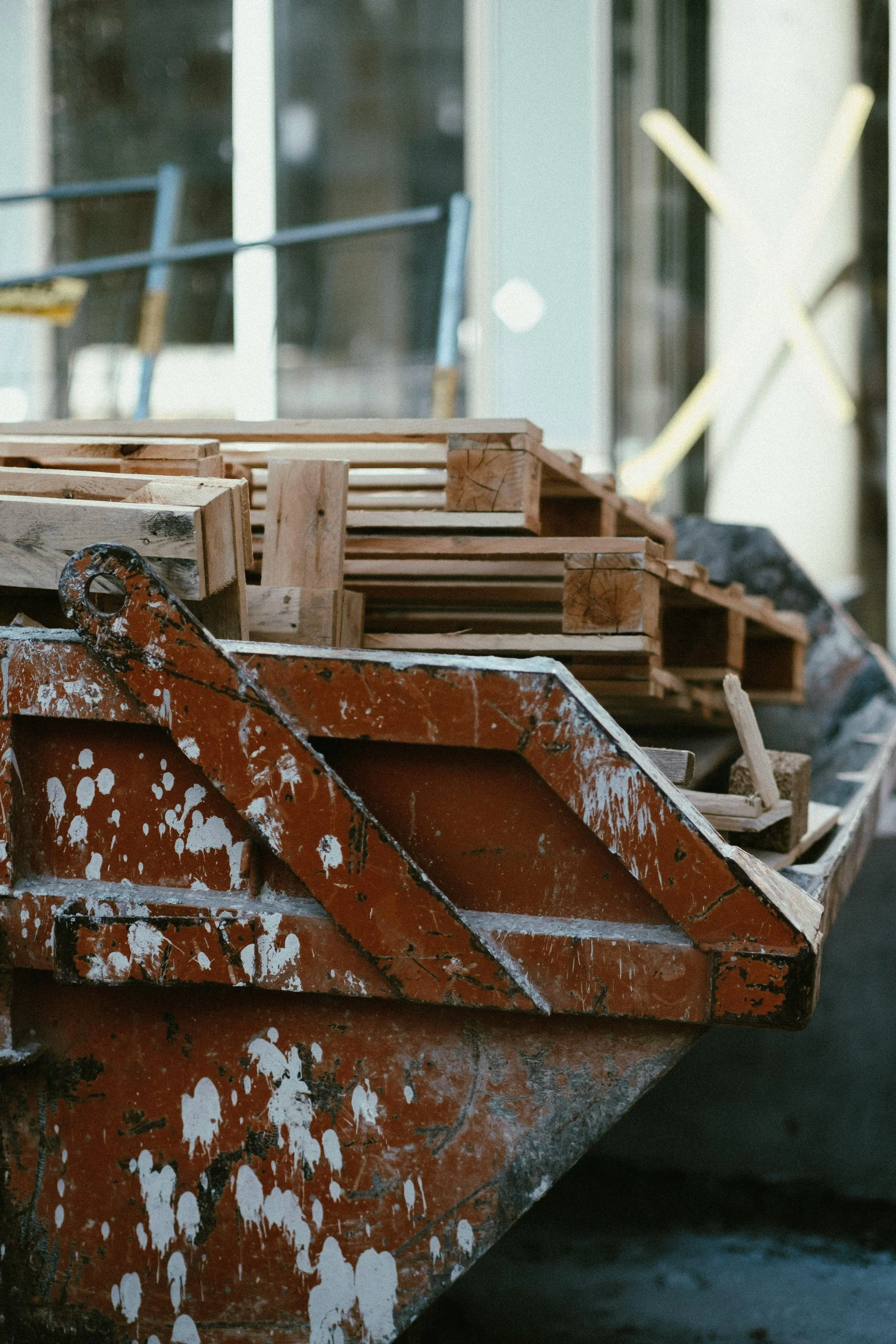Construction site with a rusted orange wheelbarrow and stacked wooden planks in front of a building with large windows.