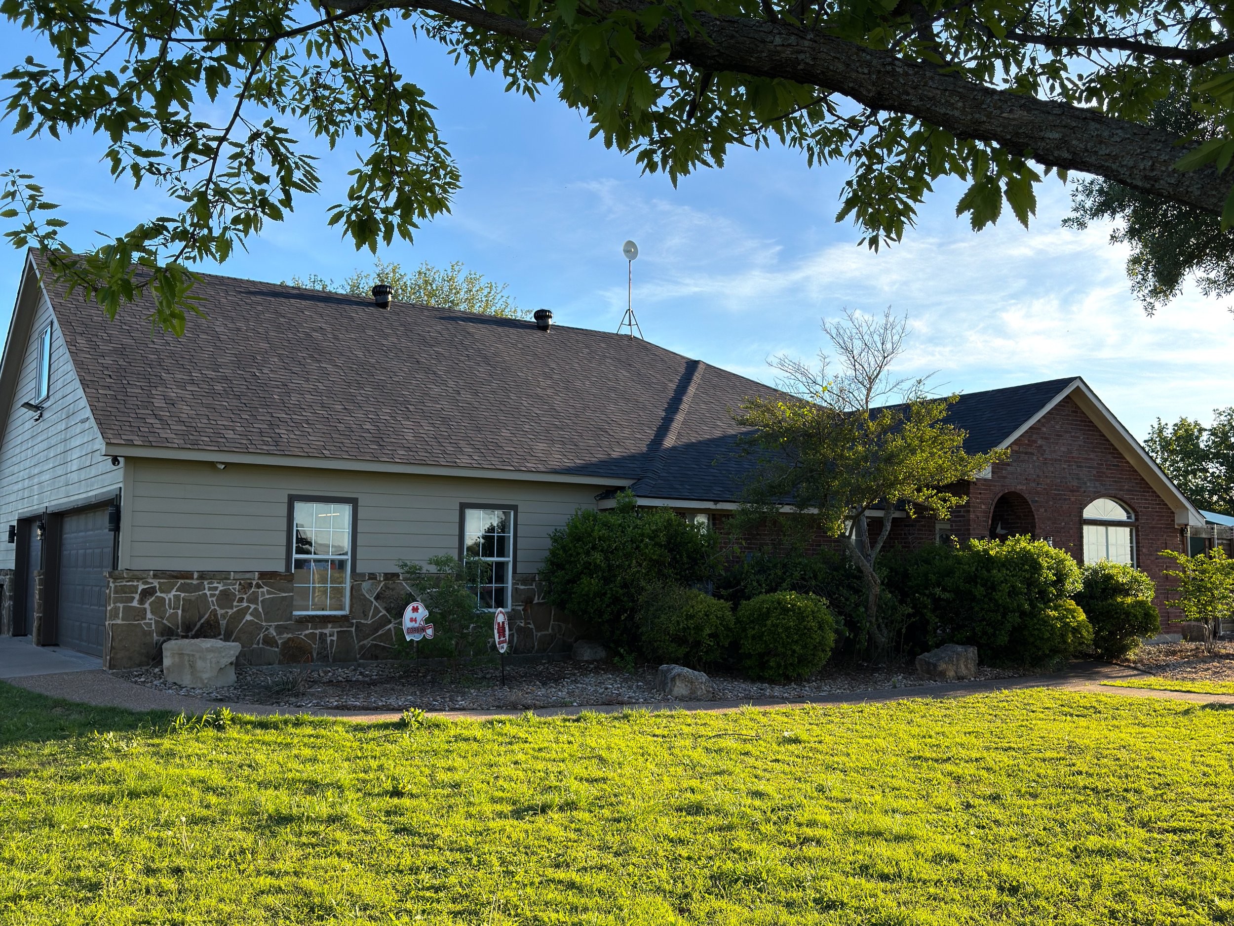 A house with a beige siding and stone exterior, dark brown roof, surrounded by green shrubs and trees, under a blue sky with some clouds, seen from a grassy yard.