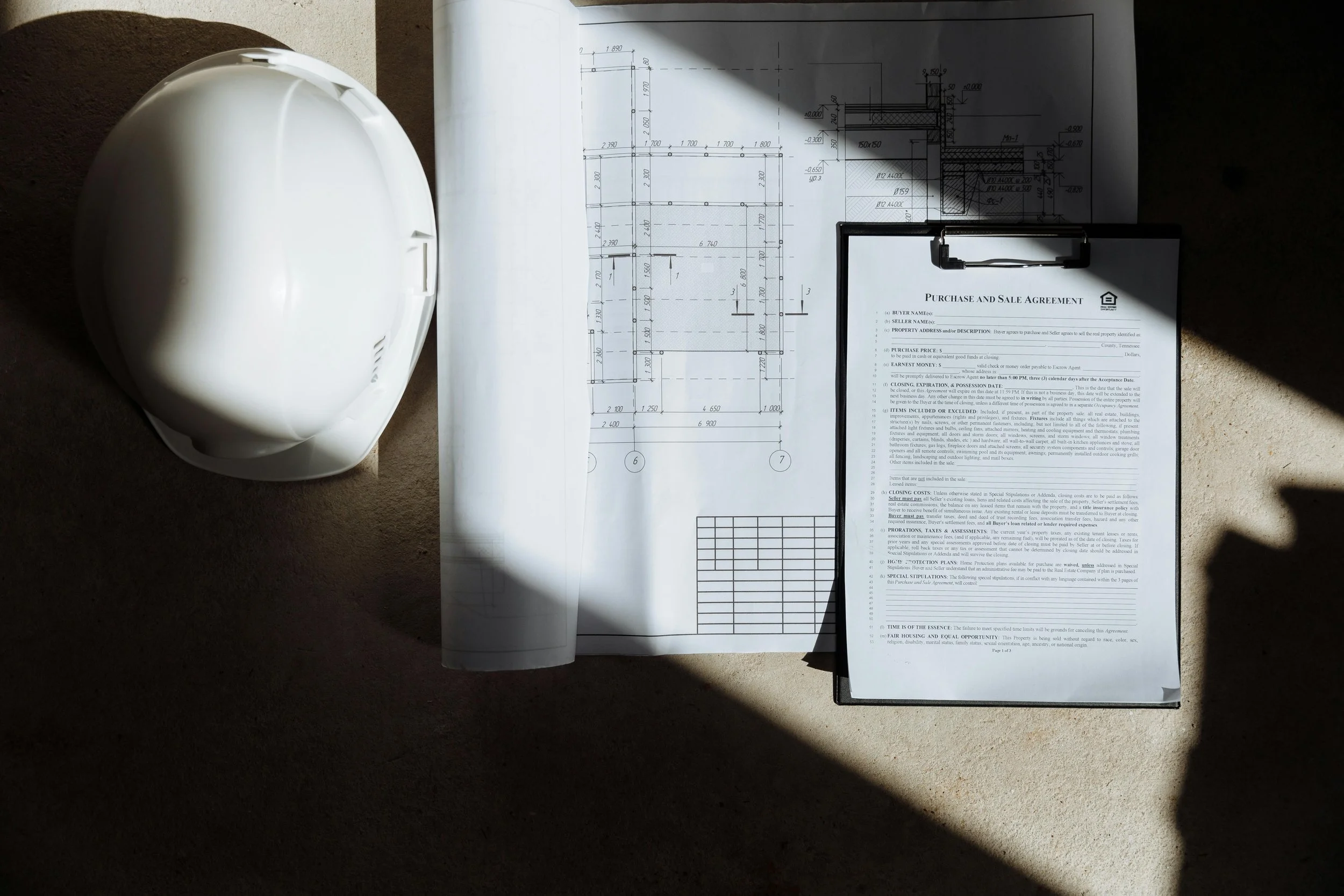 Construction helmet, architectural blueprint, and a purchase agreement on a clipboard on a concrete surface with sunlight and shadow.