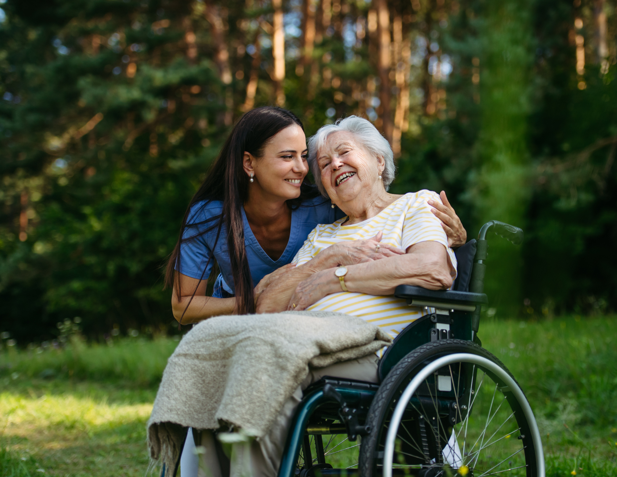 A young woman and elderly woman smiling and sharing a moment outdoors in a park, with trees in the background. The elderly woman is in a wheelchair, and they appear happy and affectionate.