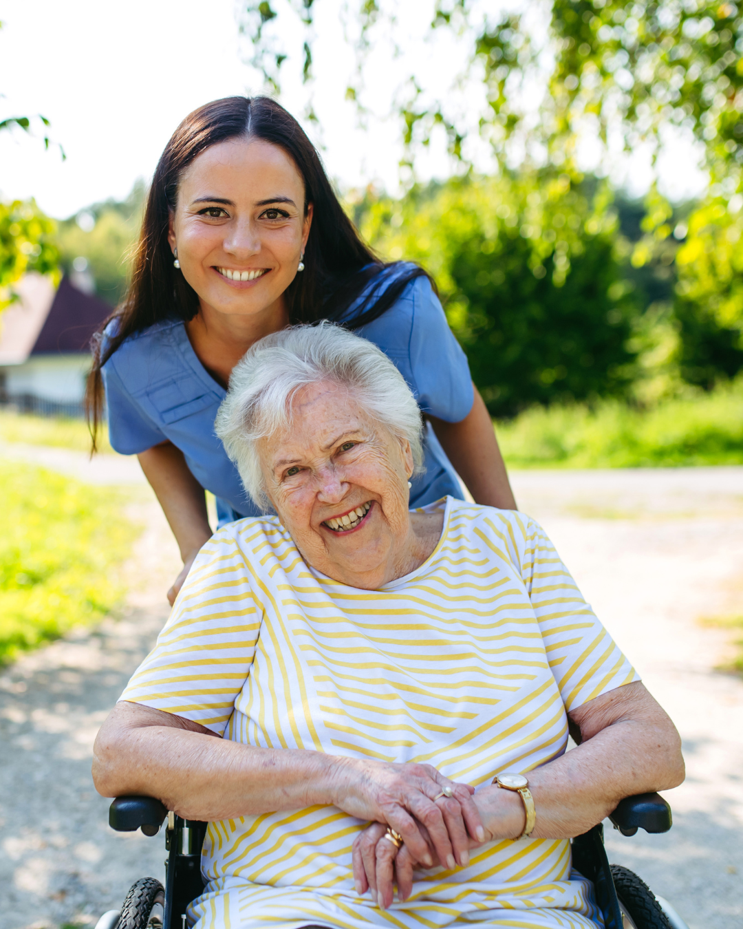 A young woman in a blue uniform leaning over an elderly woman in a wheelchair, smiling outdoors on a sunny day with green trees and a building in the background.