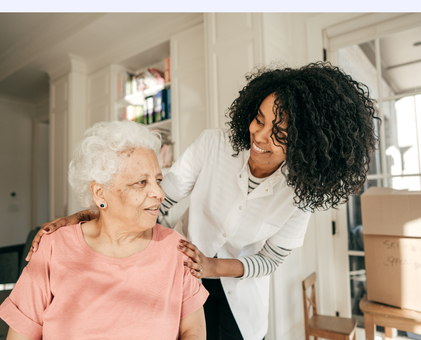 A caregiver talking to an elderly woman with white curly hair in a bright room.