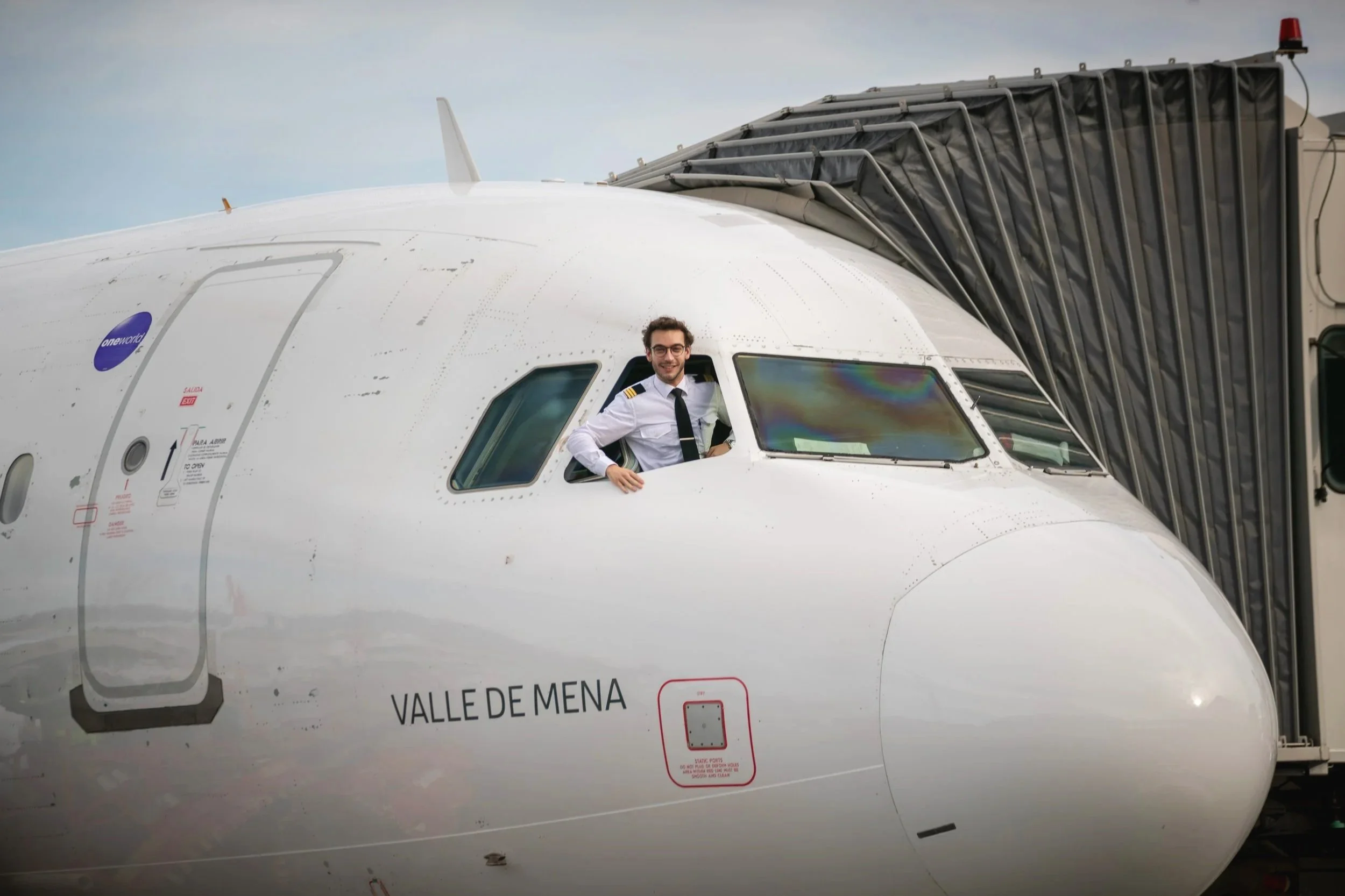A man in a pilot uniform smiling and leaning out of the cockpit of a large commercial airplane labeled 'Valle de Mena' at an airport gate.