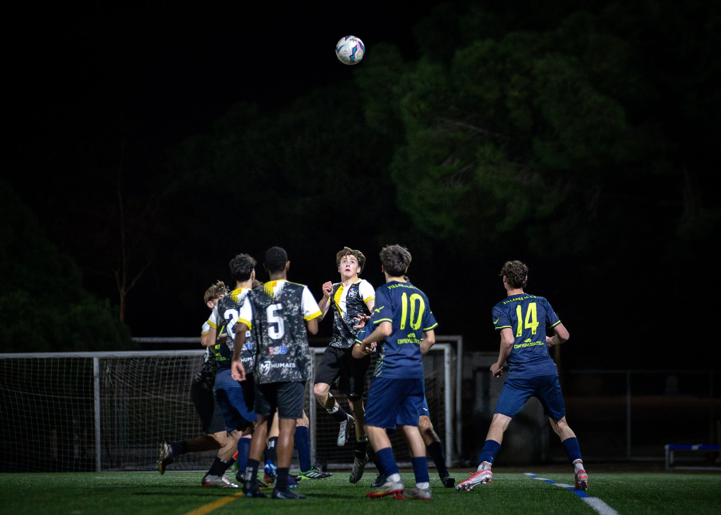 Young soccer players wearing blue and black uniforms compete for a ball on a nighttime field, with trees in the background.