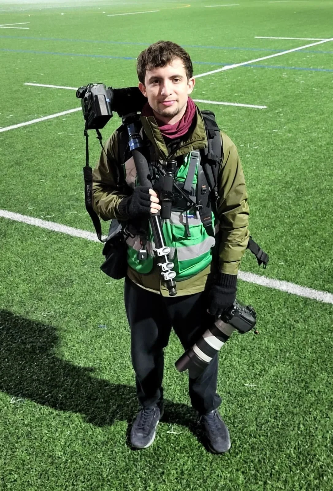 A young male photographer standing on a sports field at night, holding a camera with a large telephoto lens in his right hand and a tripod in his left. He is dressed in a green jacket, black pants, and gloves, with camera and gear strapped around his body and backpack.