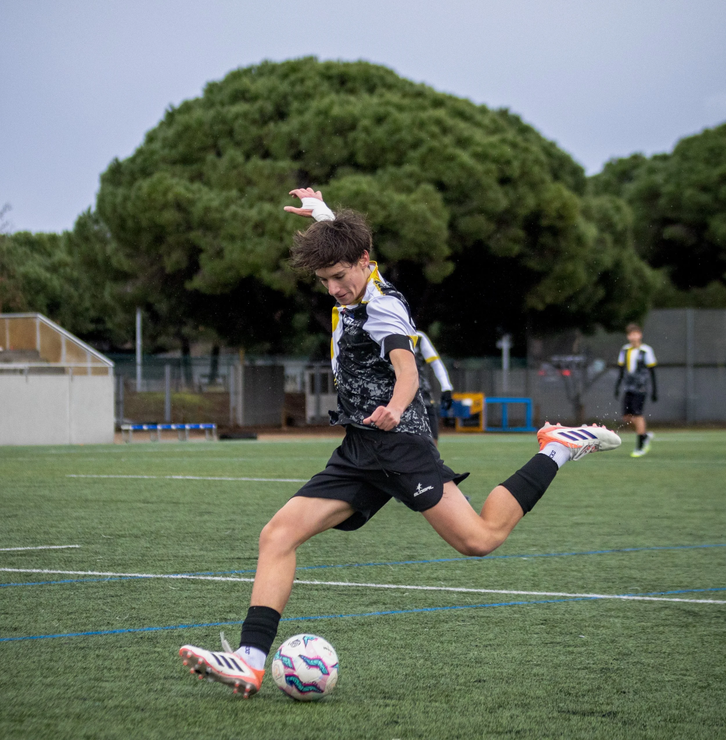 A young male soccer player in black and white kit kicking a colorful soccer ball on a synthetic field, with trees and other players in the background during evening.