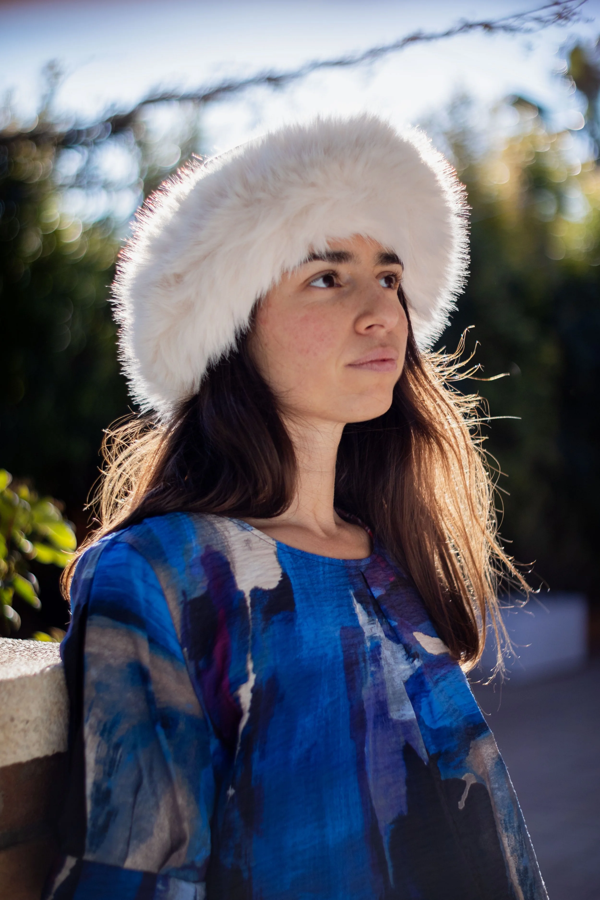 A young woman wearing a fluffy white hat and a blue patterned top, sitting outdoors with blurred greenery and sunlight in the background.