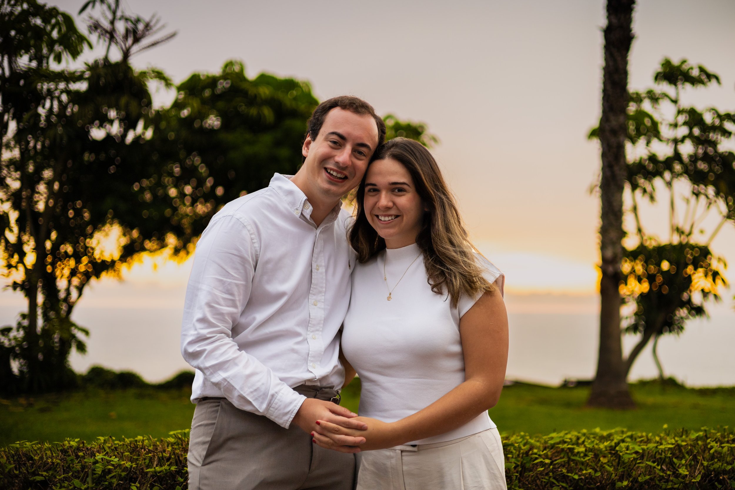 A smiling couple, a man and a woman, standing close together outdoors during sunset, holding hands, with trees and a body of water in the background.