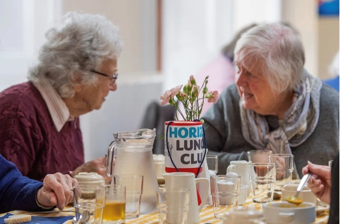 Two elderly women smiling and talking at a lunch table with glasses, a pitcher, and a flower centerpiece.