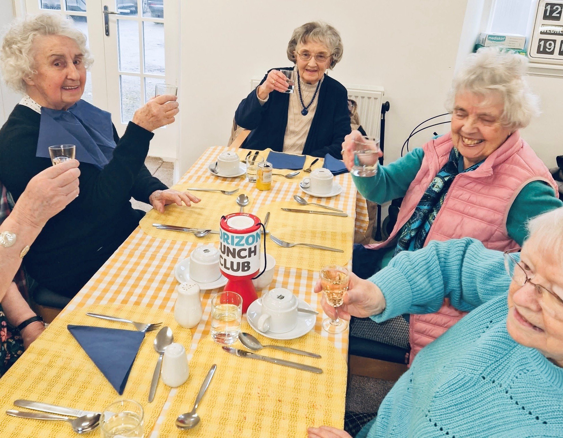 Group of five elderly women sitting around a table with drinks, raising glasses for a toast, in a bright room with a yellow checkered tablecloth.