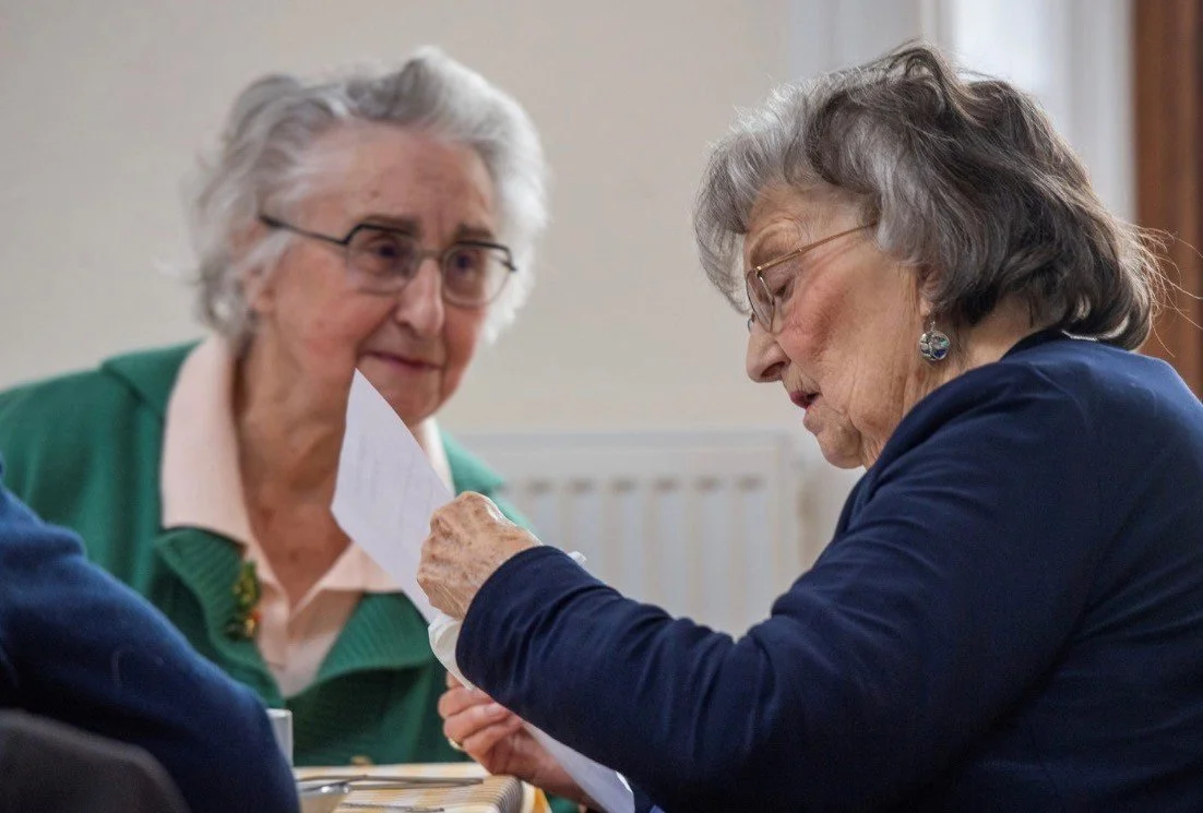 Two elderly women, one wearing glasses and a blue blazer, the other with white hair and glasses, sitting closely and looking at a paper the woman in the blazer is holding.