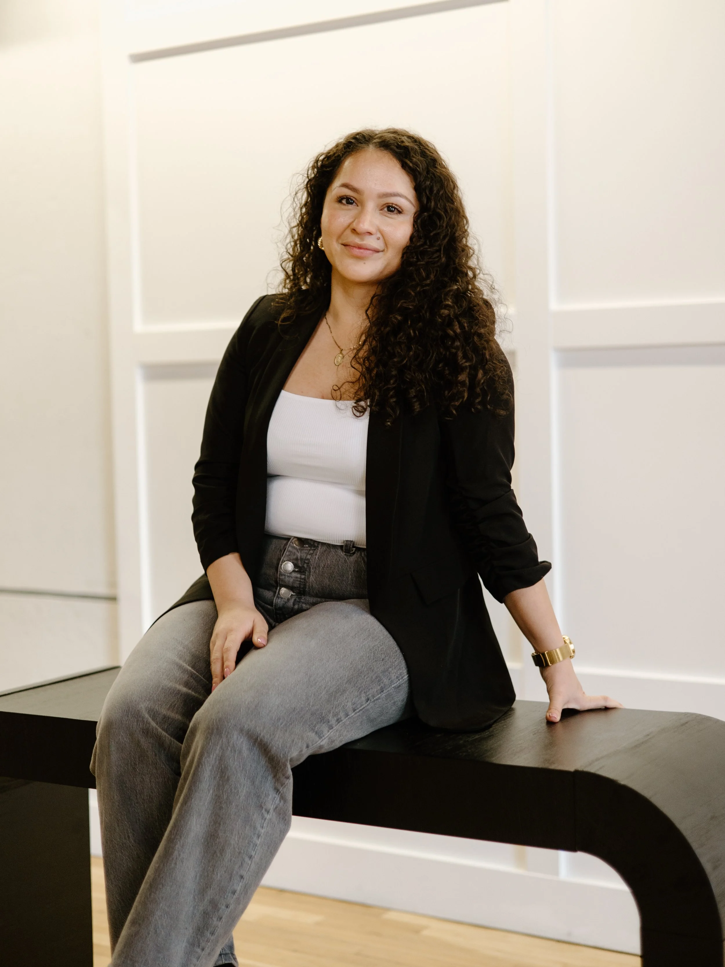 A young woman with curly brown hair, wearing a black blazer, white top, and gray jeans, sitting on a black bench in a modern indoor setting with white walls.