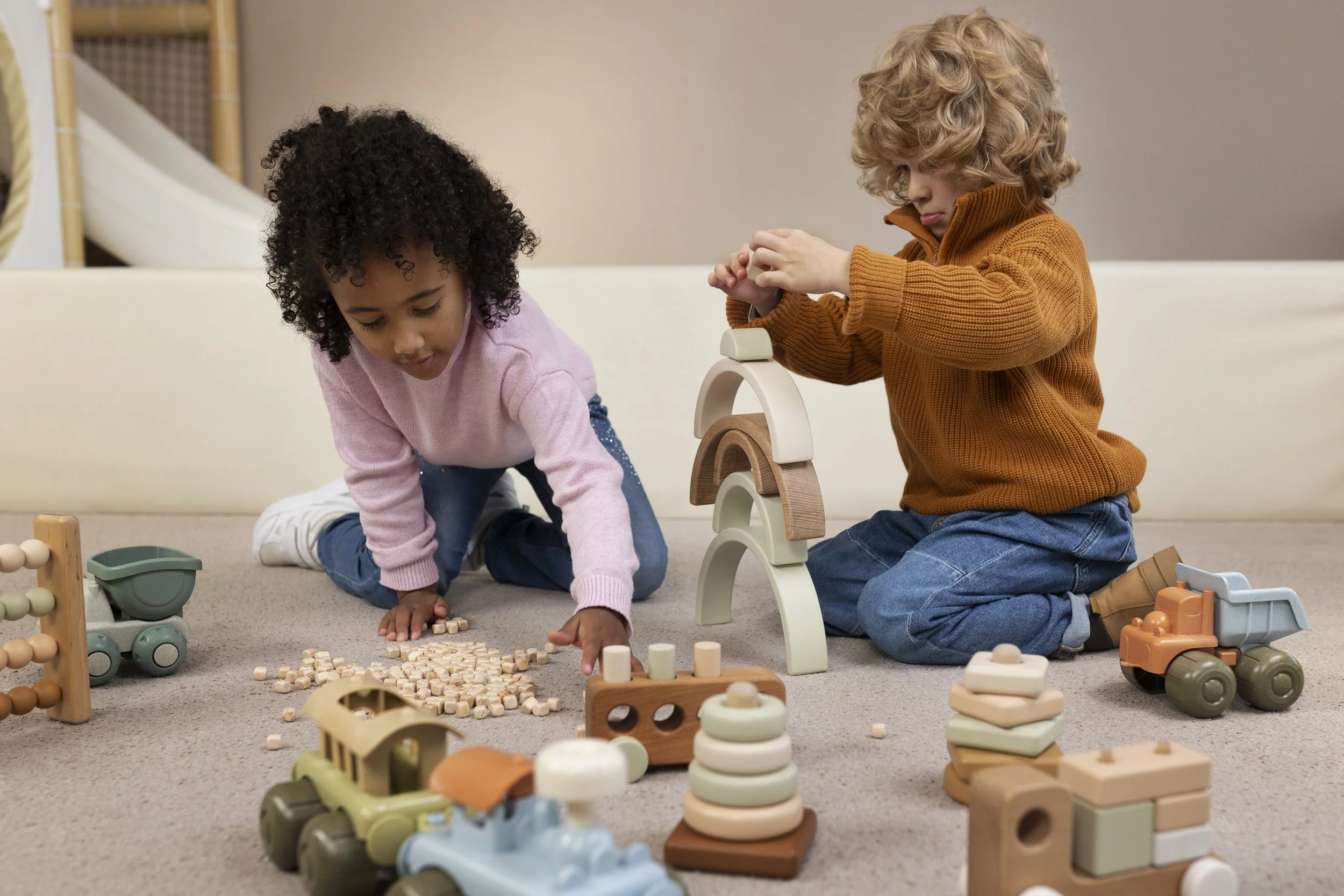 Two children playing on the carpeted floor with wooden toys, including stacking blocks, vehicles, and an abacus, in a cozy room.