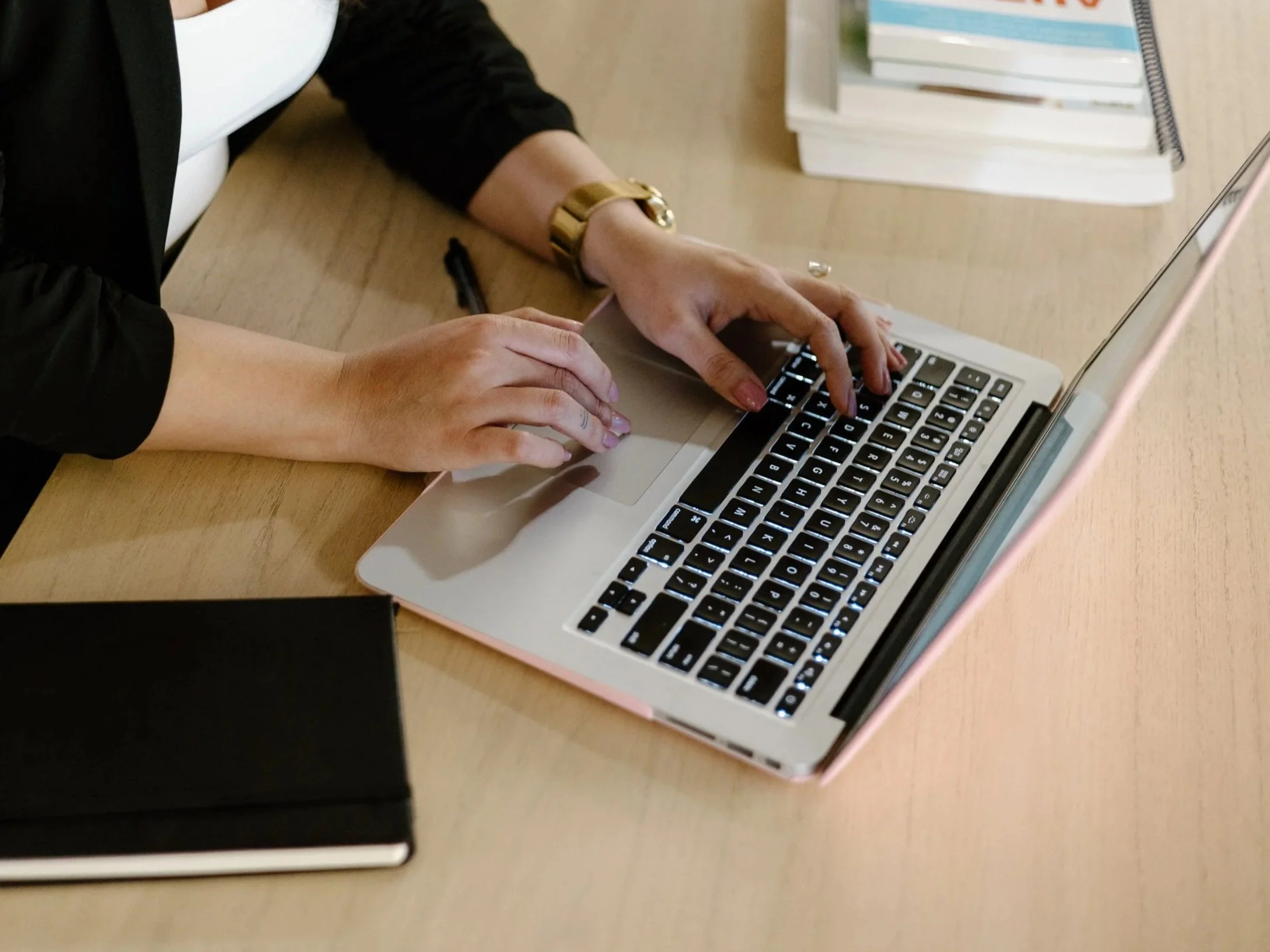 Close-up of a person typing on a silver laptop with black keys, on a wooden desk next to a stack of books and a black notebook.
