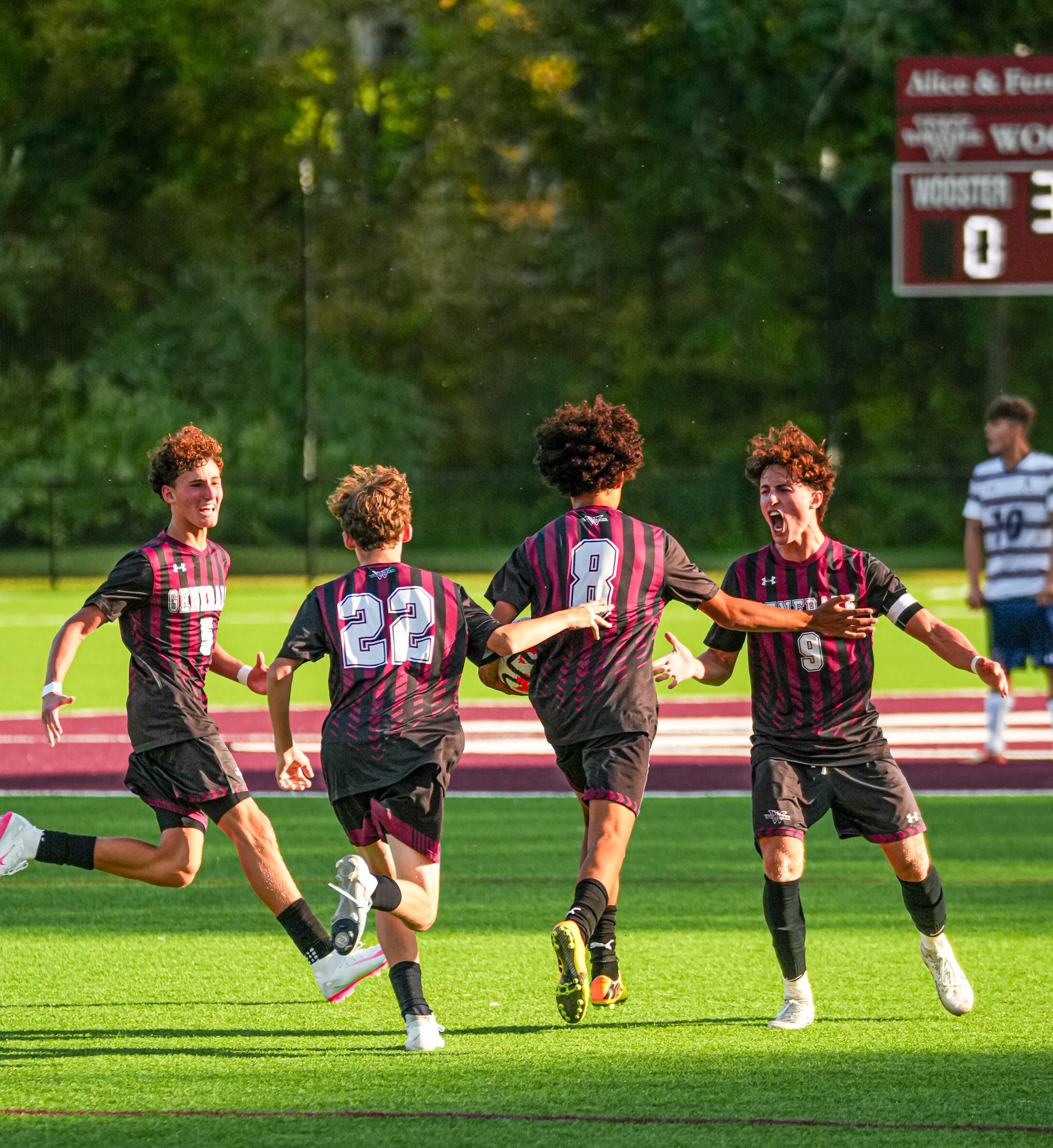 Soccer players celebrating on the field, wearing black and maroon uniforms, with a scoreboard in the background.