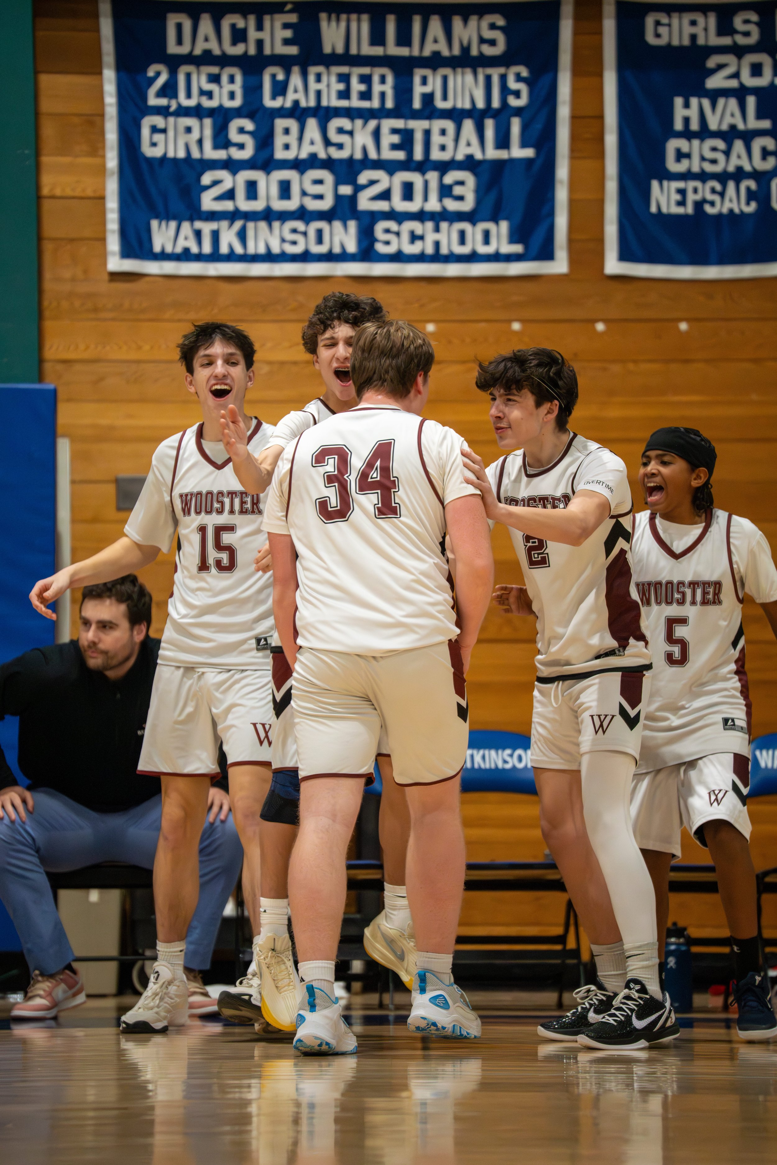 A group of young basketball players in white uniforms celebrating on the court, with a coach sitting beside them, in a gymnasium with wooden walls and banners in the background.