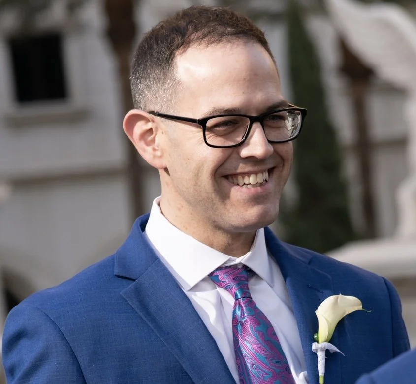 A man dressed in a blue suit, white shirt, and colorful purple and blue tie, smiling outdoors. He has short brown hair, glasses, and a white flower boutonniere on his lapel.