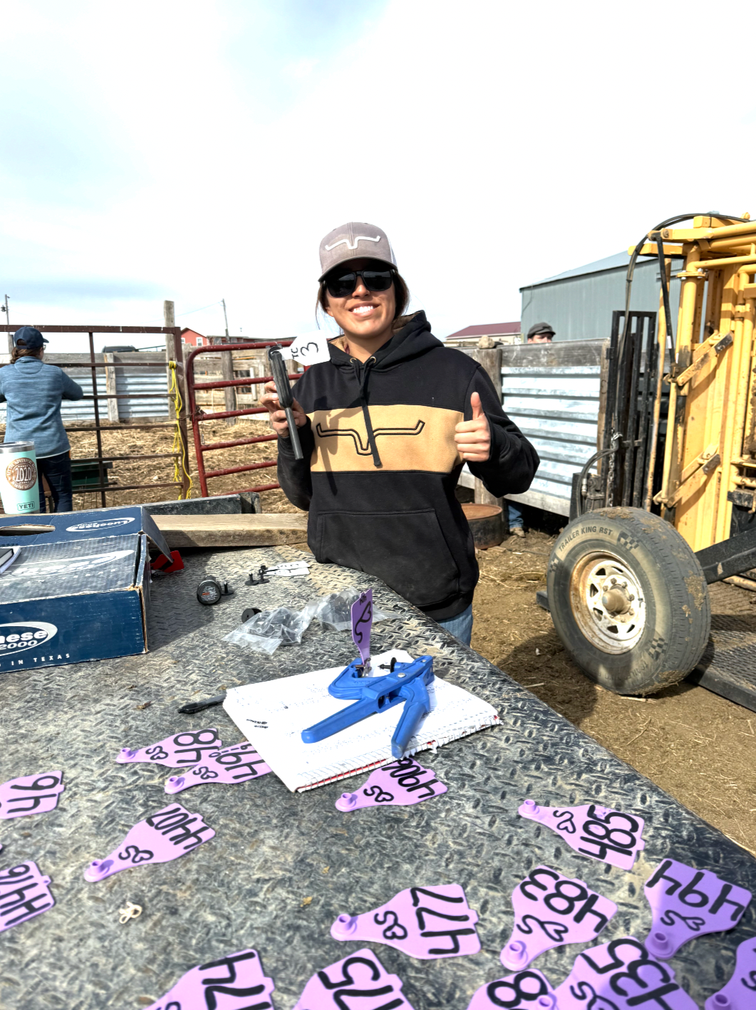 A woman wearing sunglasses, a gray cap, and a black hoodie with tan stripes is smiling and giving a thumbs-up at an outdoor event, likely on a farm or ranch. She stands behind a table with pink numbered tags, a cardboard box, and a blue tool on it. There are other people and farm equipment in the background.