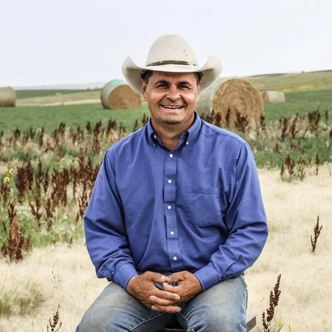 A man smiling and sitting outdoors in a field with hay bales in the background. He is wearing a cowboy hat and a blue button-up shirt.