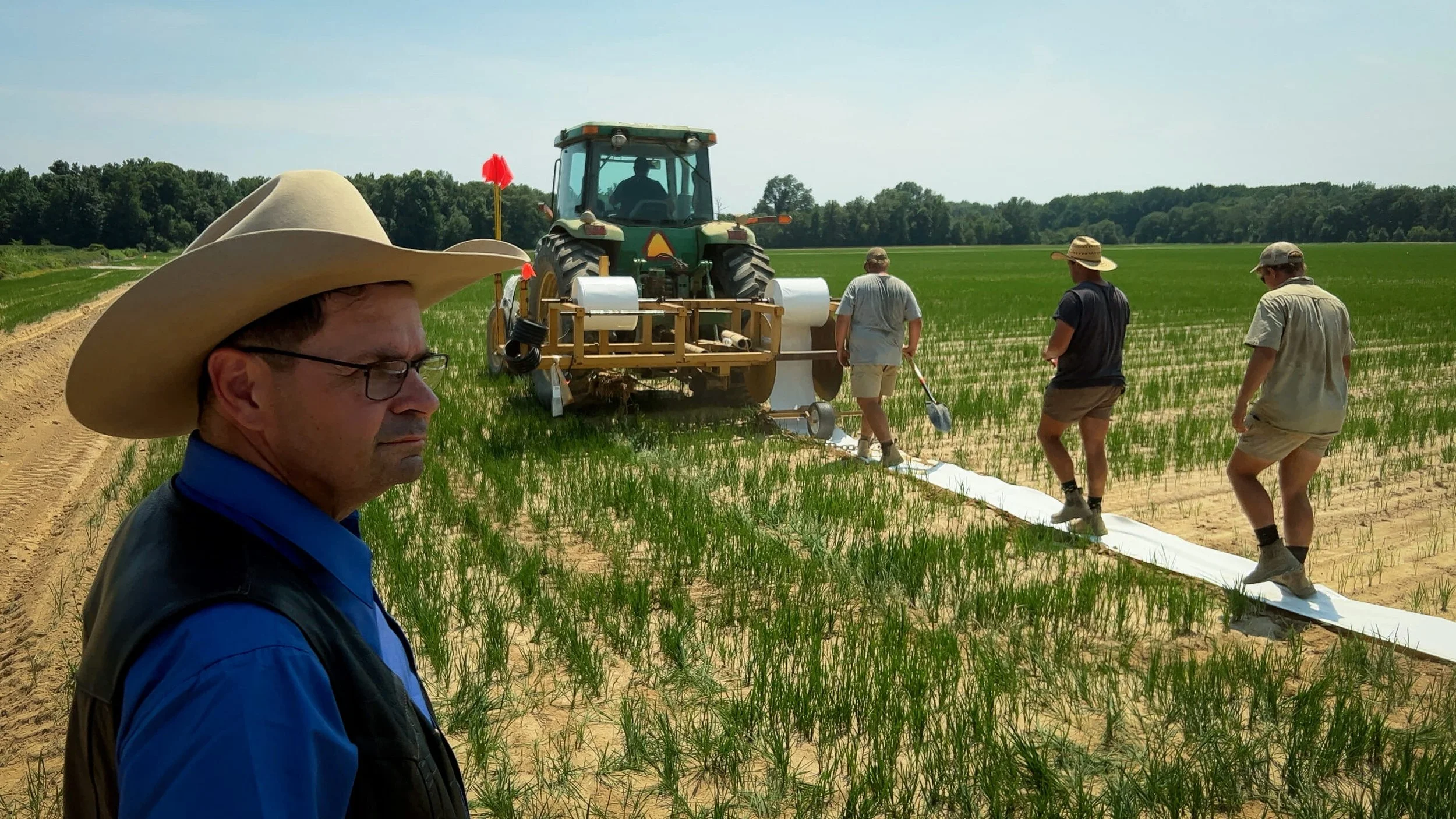 A group of people working in a field planting crops with a tractor and planting equipment, with a man in a cowboy hat in the foreground.