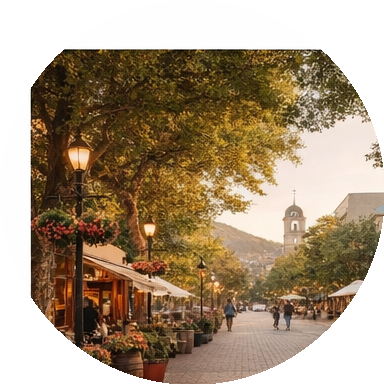 A lively outdoor street scene during fall with trees and street lamps lining a pedestrian walkway. There are people walking, storefronts with umbrellas, and a prominent clock tower in the background.